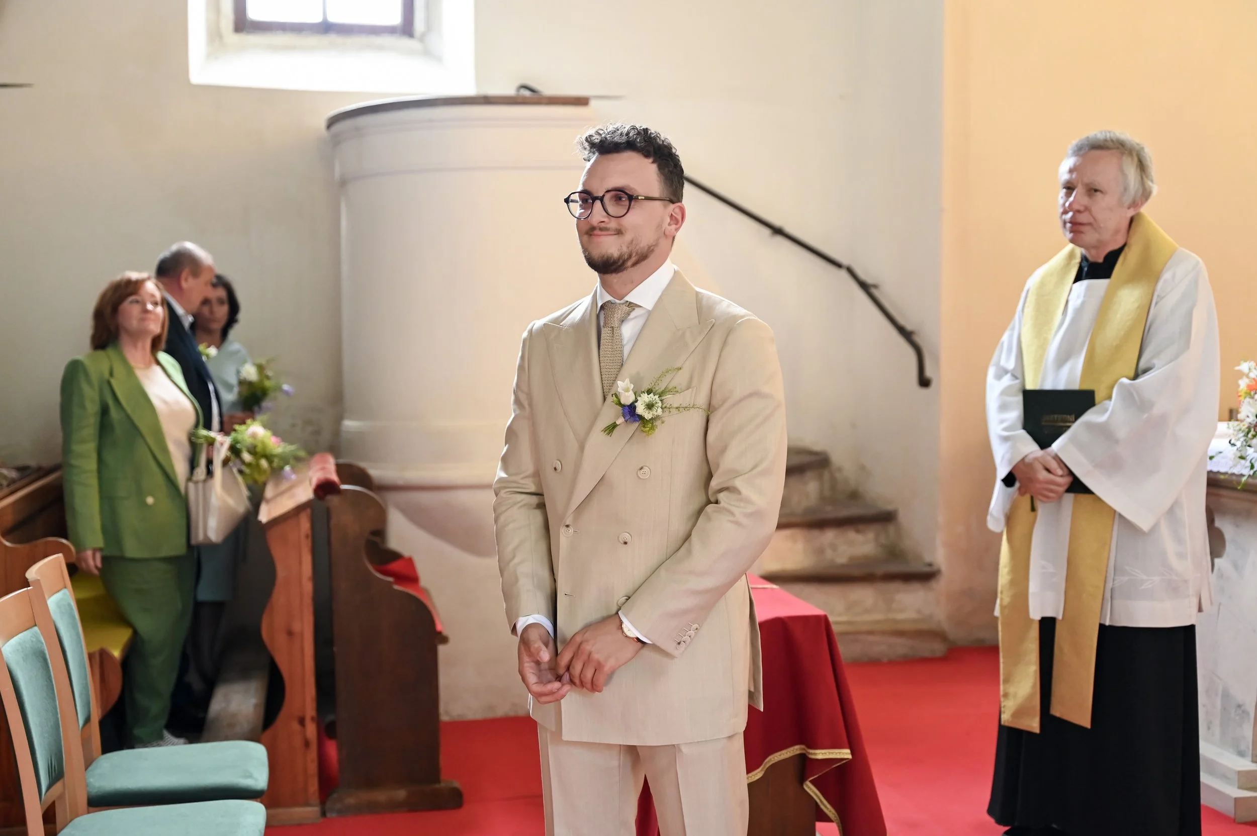 Young man in cream-colored suit with glasses and boutonniere standing in a church during a wedding ceremony.