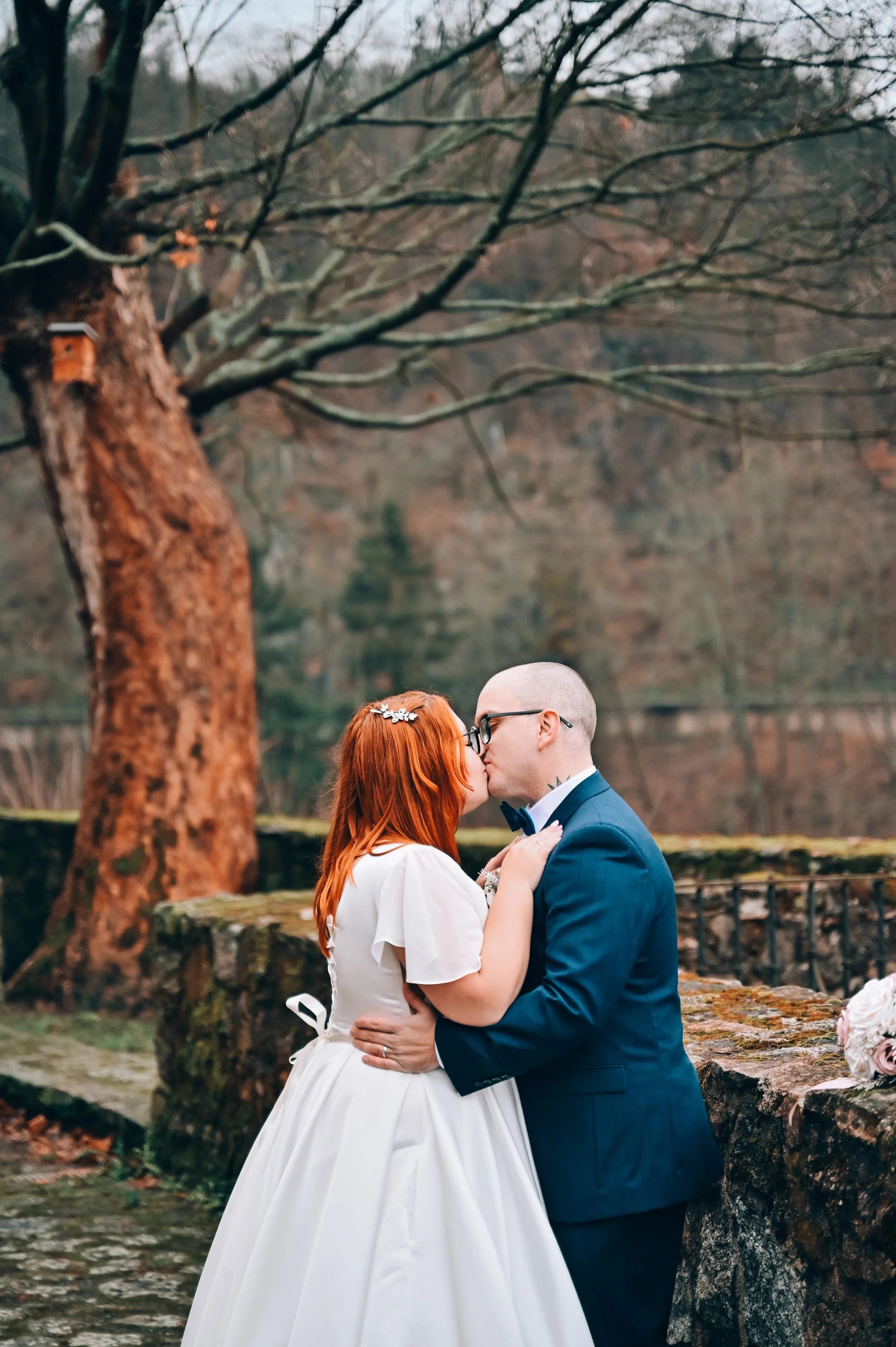 A couple dressed in wedding attire sharing a kiss outdoors in front of a large tree and stone wall.