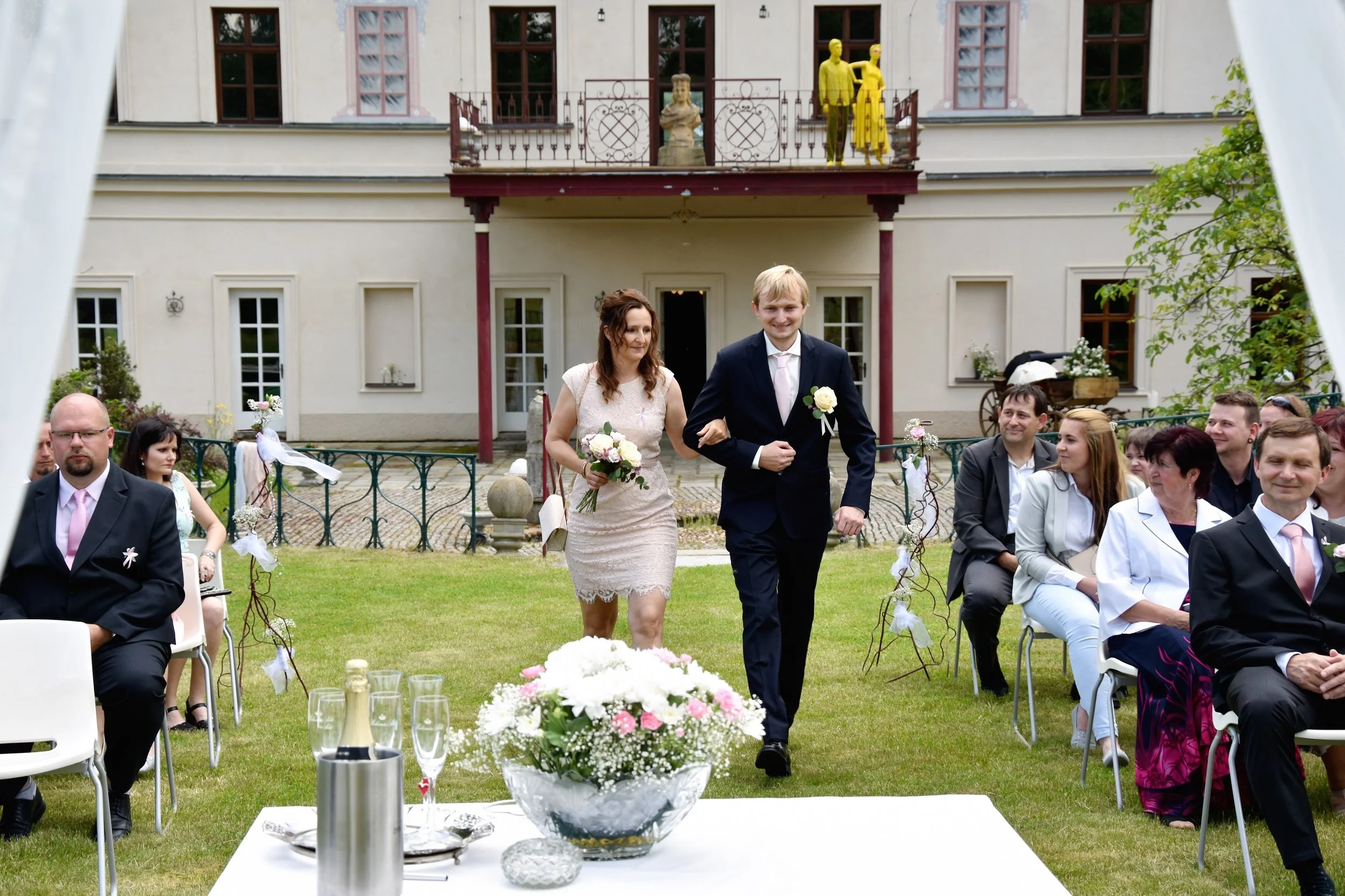 Nevěsta a ženich kráčejí uličkou na svatebním obřadu venku, zatímco je sledují sedící hosté.