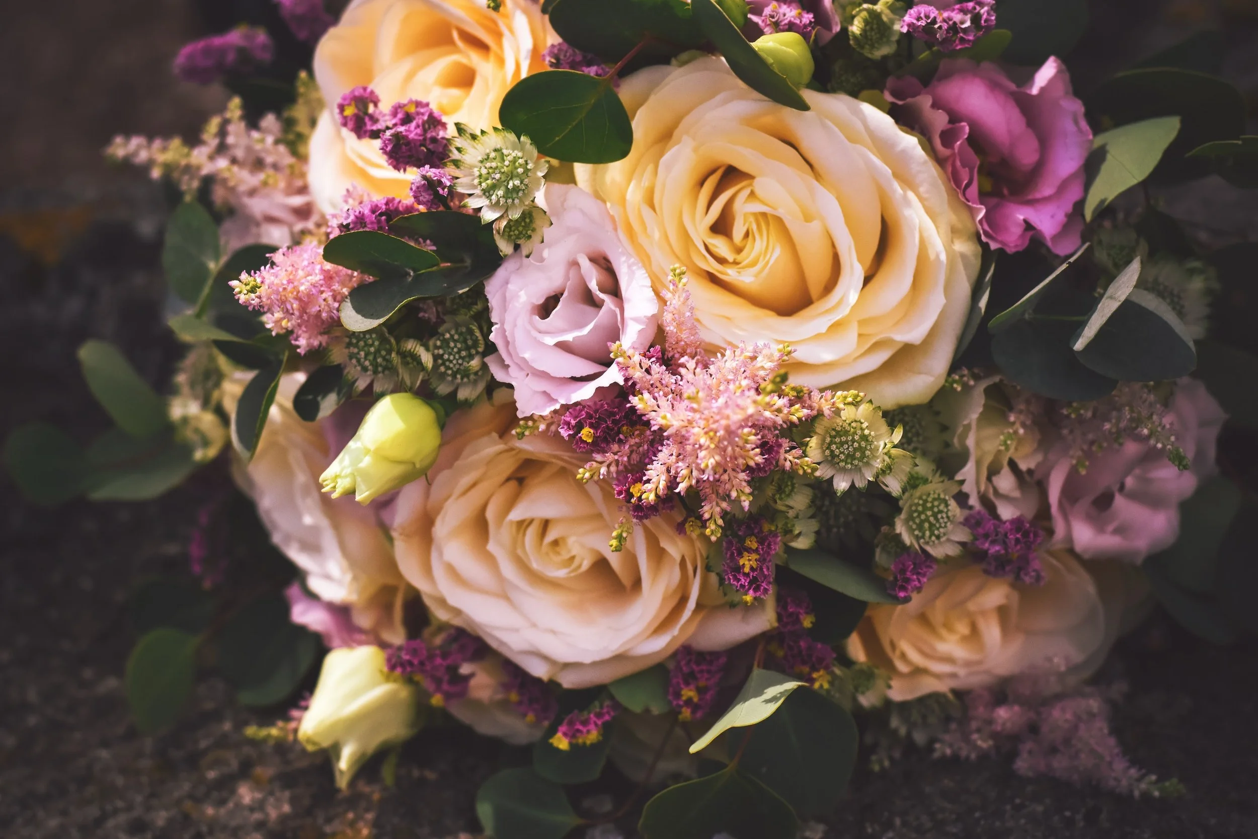 Kytice krémově zbarvených růží, růžového lisianthusu, fialových květů, zelených listů a malých bílých a fialových květů.