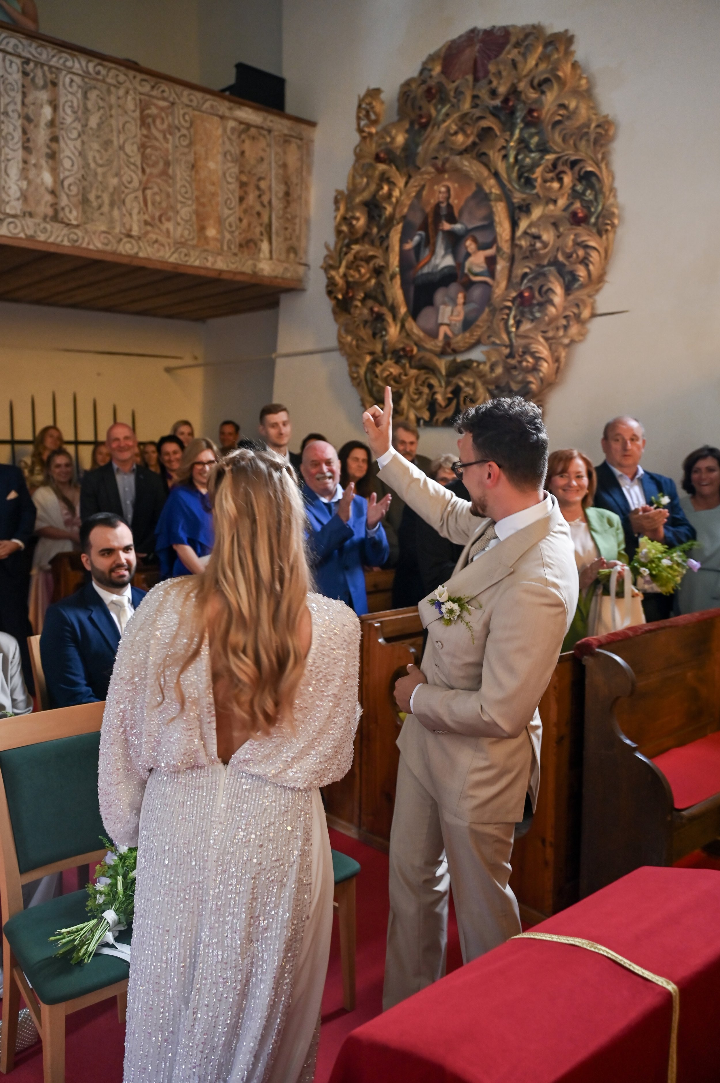 A couple is exchanging vows during a wedding ceremony in a church, with the officiant raising one hand. The bride has long, wavy blonde hair and wears a glittering, open-back wedding dress. The groom wears a beige suit. Guests in formal attire watch 