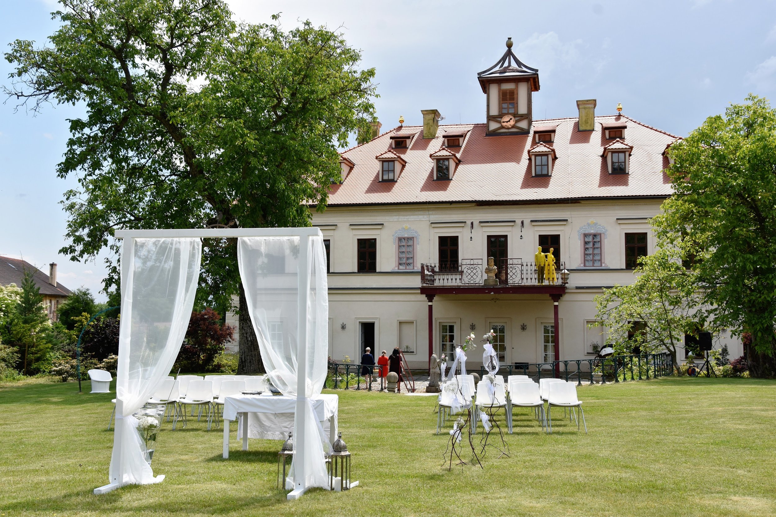 Venkovní svatební scenérie s bílým obloukem a židlemi na trávníku před velkým historickým domem s červenou střechou a hodinovou věží. Dekorace zahrnují bílou látku a lucerny. Zámek Rochlov