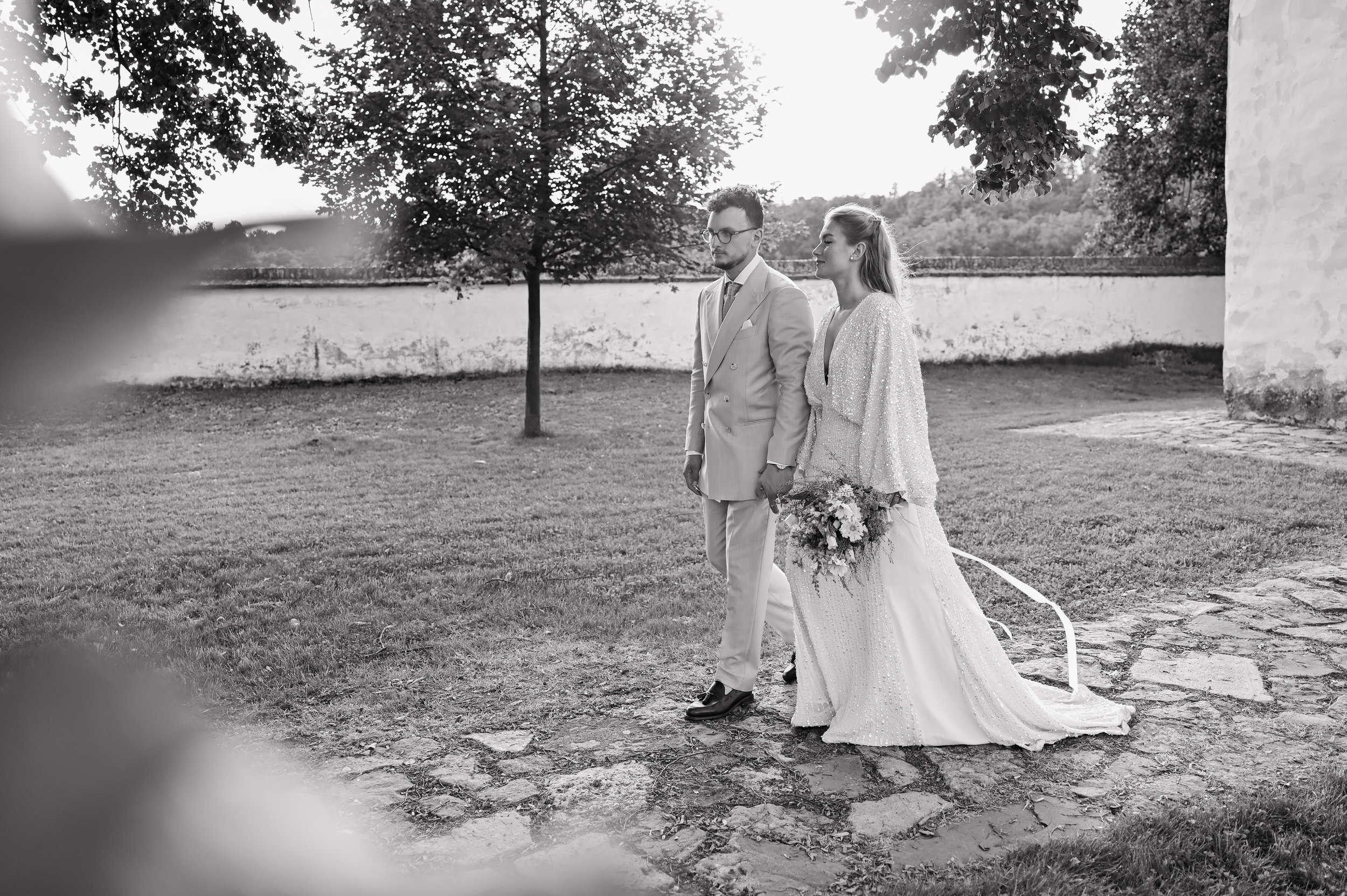 Black and white photo of a bride and groom walking outdoors, with the bride holding a bouquet, near a stone path and trees.