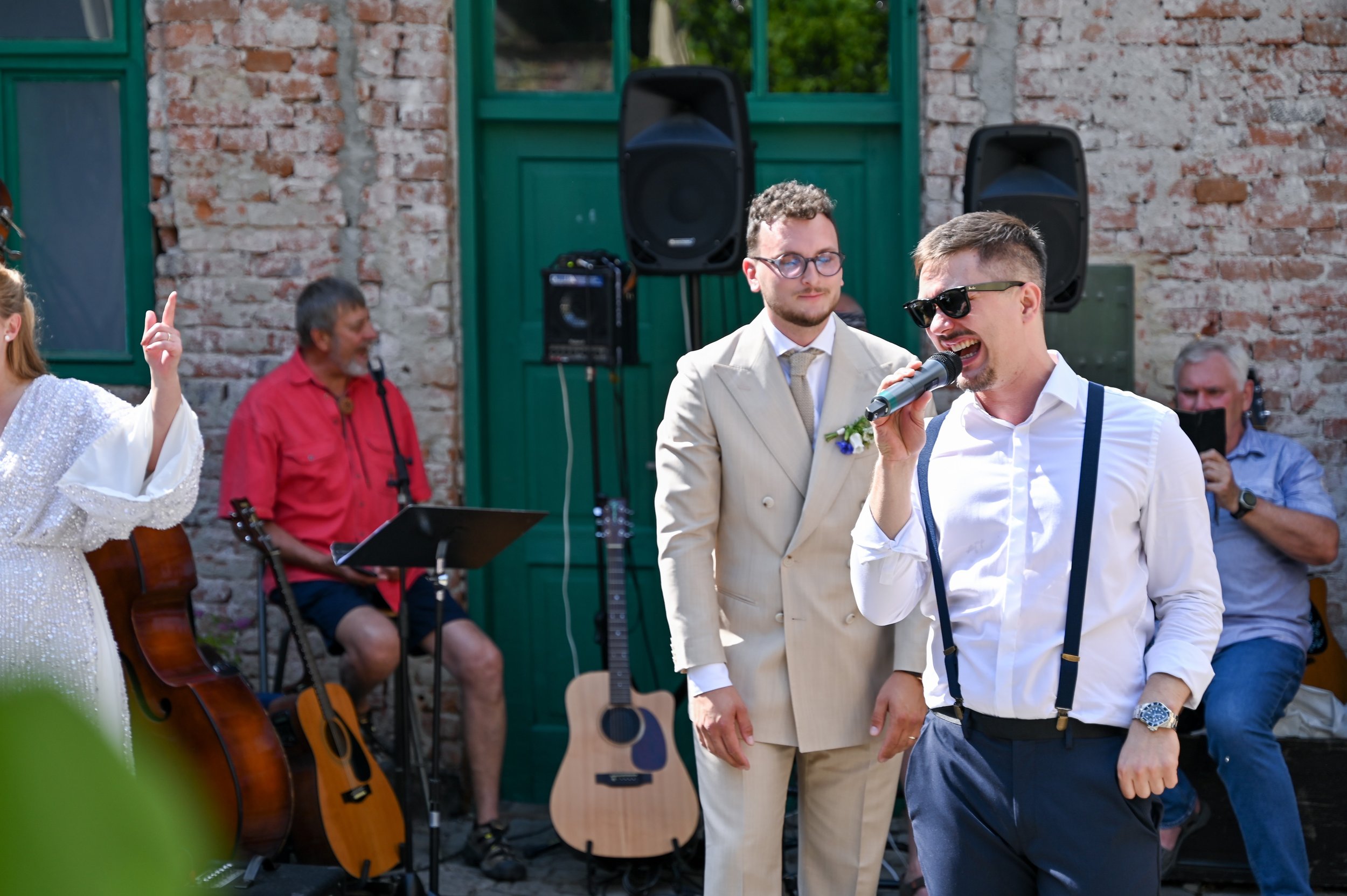 A man in sunglasses singing into a microphone at an outdoor gathering, with a man in a beige suit and glasses standing behind him, and musicians with guitars in the background against a brick wall.