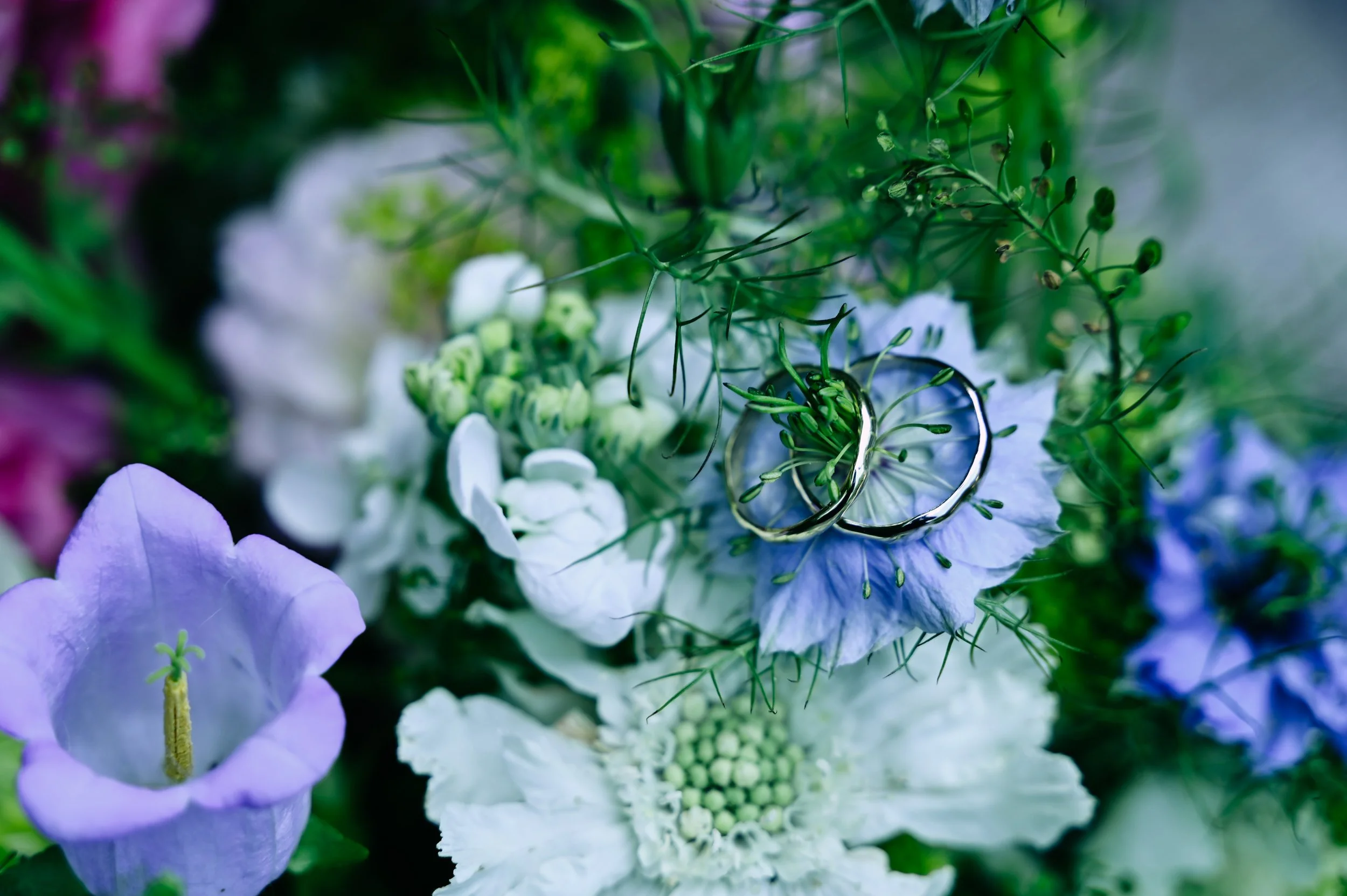 Close-up of a floral arrangement with white, purple, and blue flowers, decorated with two silver wedding bands.