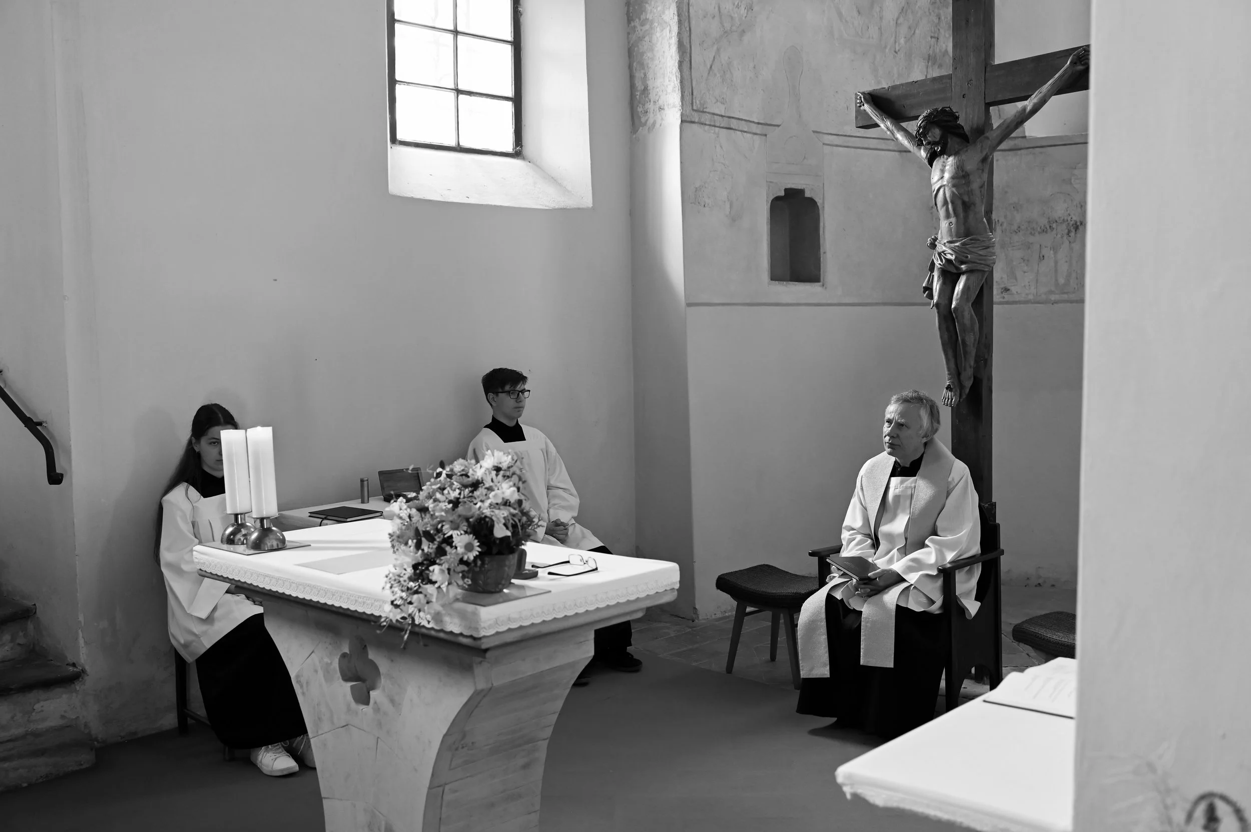 A black and white photo of a religious ceremony inside a church. A priest sitting on a chair with a book, two altar servers sitting behind the altar, and a large crucifix with Jesus Christ on the cross on the wall.