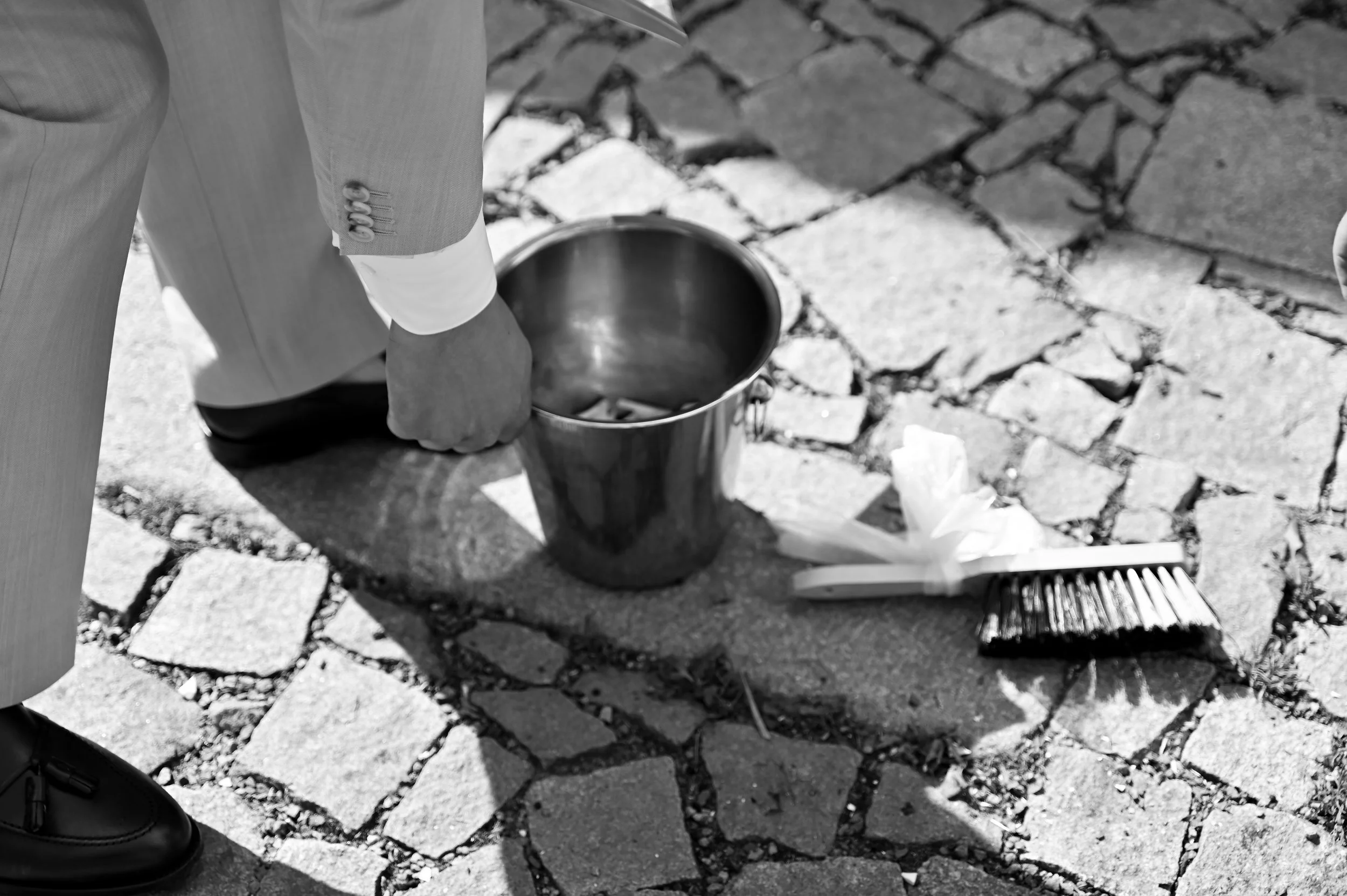 A person in a suit is cleaning a cobblestone street with a small broom and dustpan, with a metal bucket nearby. The person’s hand is reaching into the bucket, and they are wearing black shoes.