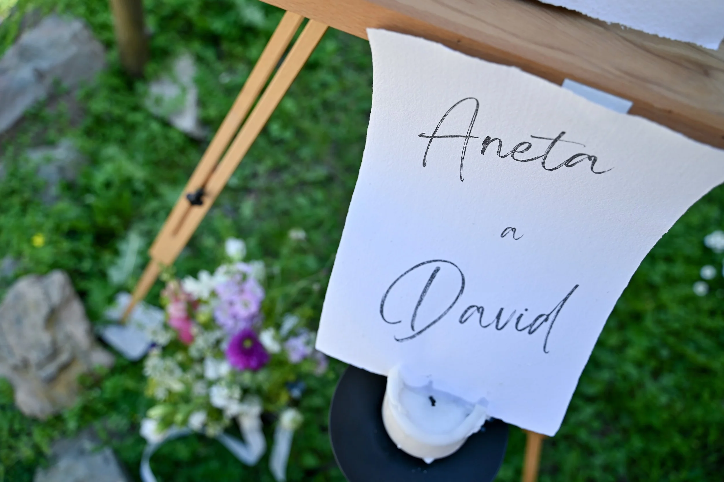 A handwritten sign labeled 'Aneta a David' attached to a black candle holder at an outdoor setting, with a flower arrangement and a wooden structure in the background.
