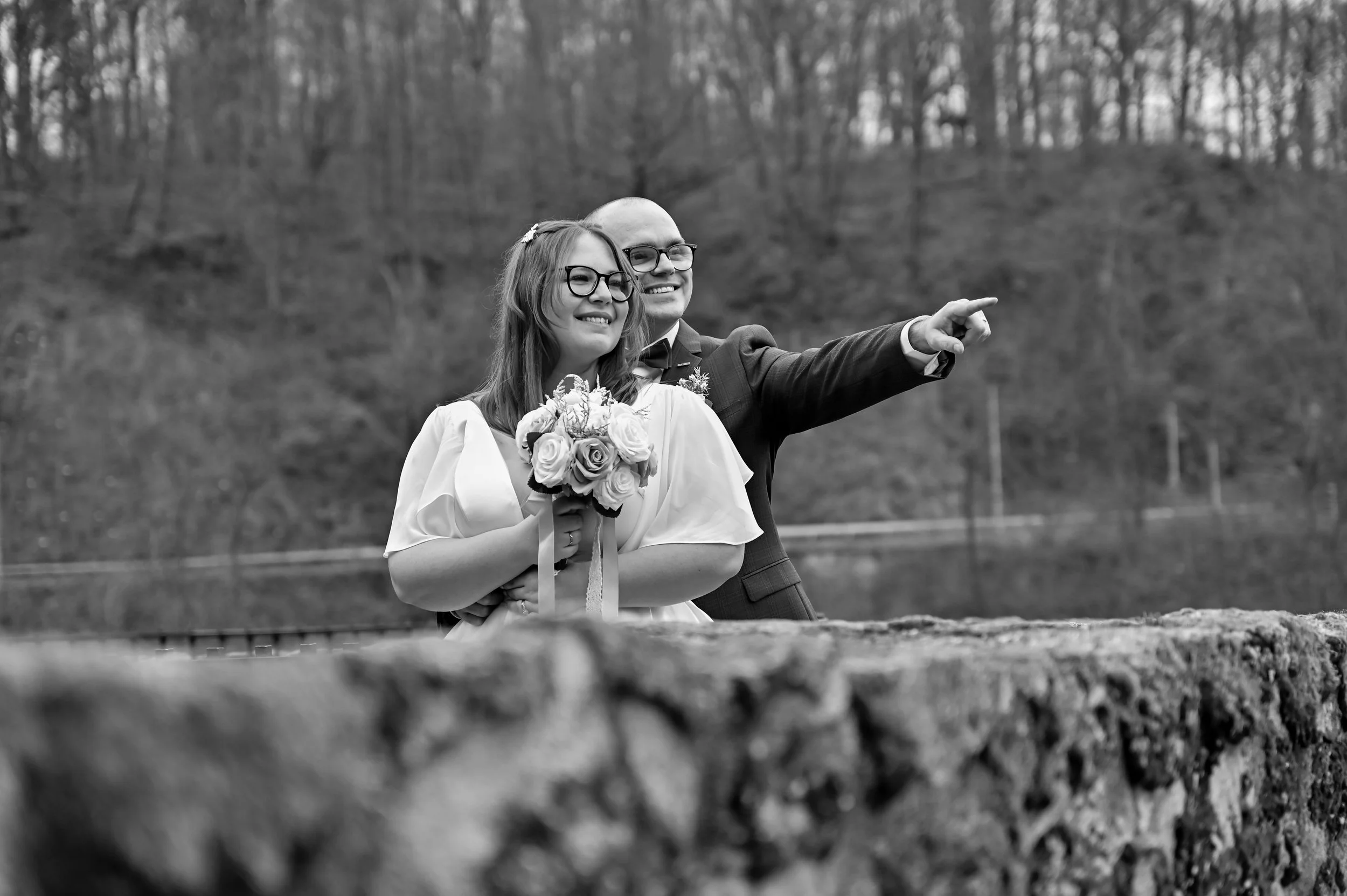 A black-and-white photograph of a wedding couple outdoors, with a woman holding a bouquet of roses, standing beside a man in a suit who is pointing at something in the distance, with a natural landscape of trees and a hillside in the background.