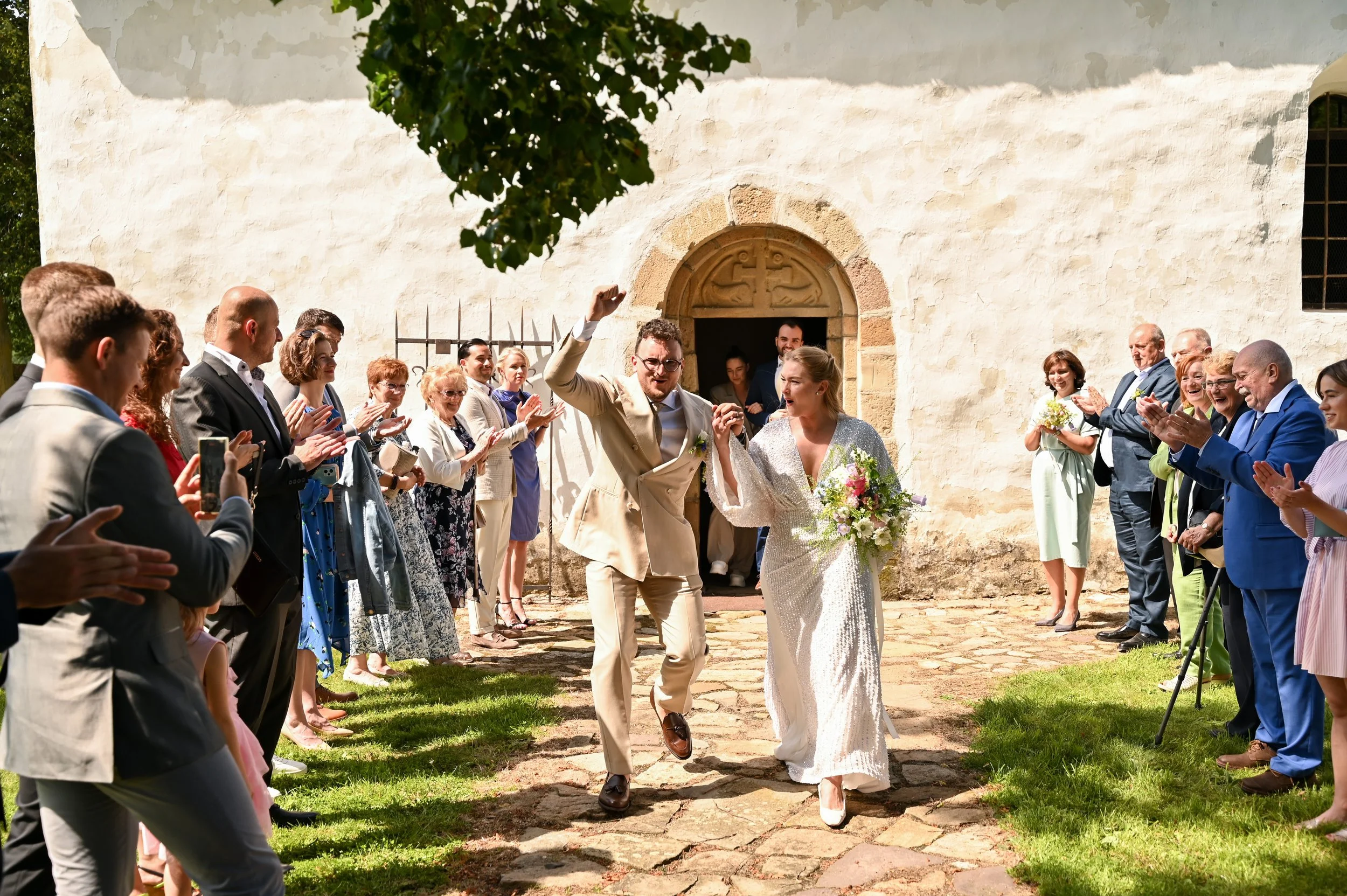A newlywed couple celebrating as they exit a church, surrounded by guests clapping and taking photos on a sunny day.