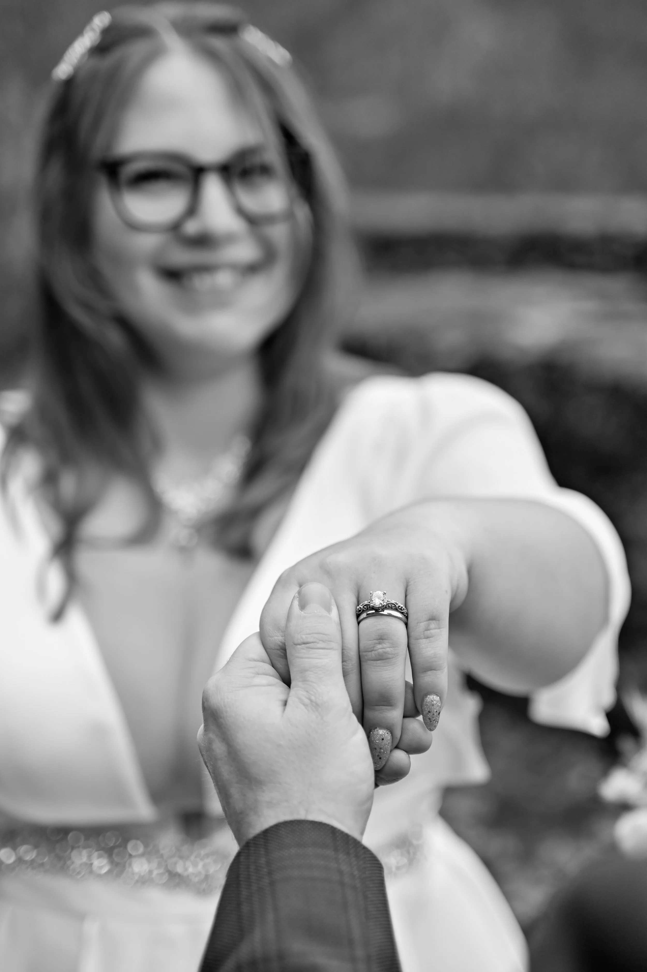 A woman with glasses smiling, holding her hand with an engagement ring on her finger, in focus, black and white photo.