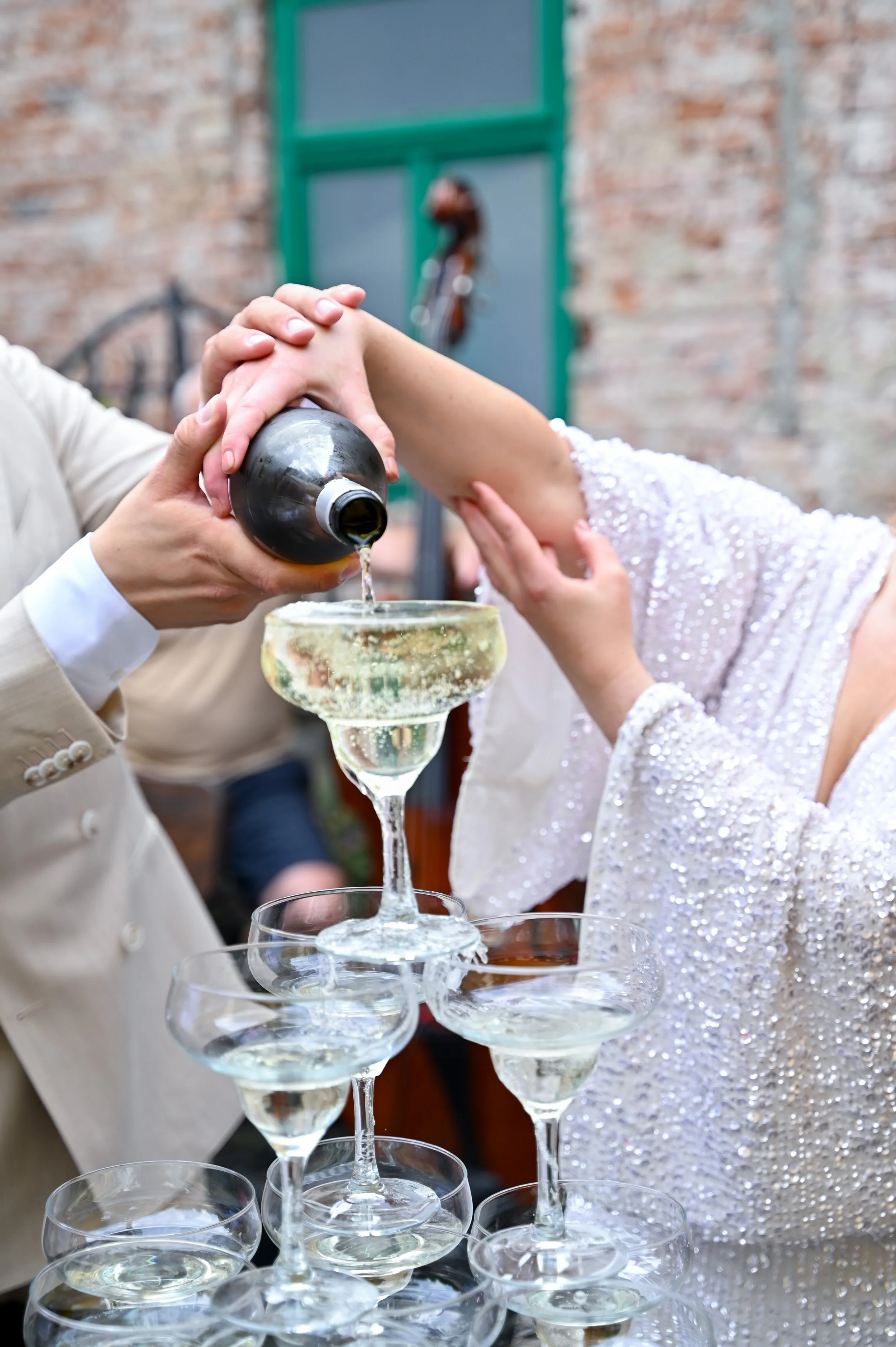People pouring champagne into a pyramid of glasses during a celebration, with a brick wall and window in the background.