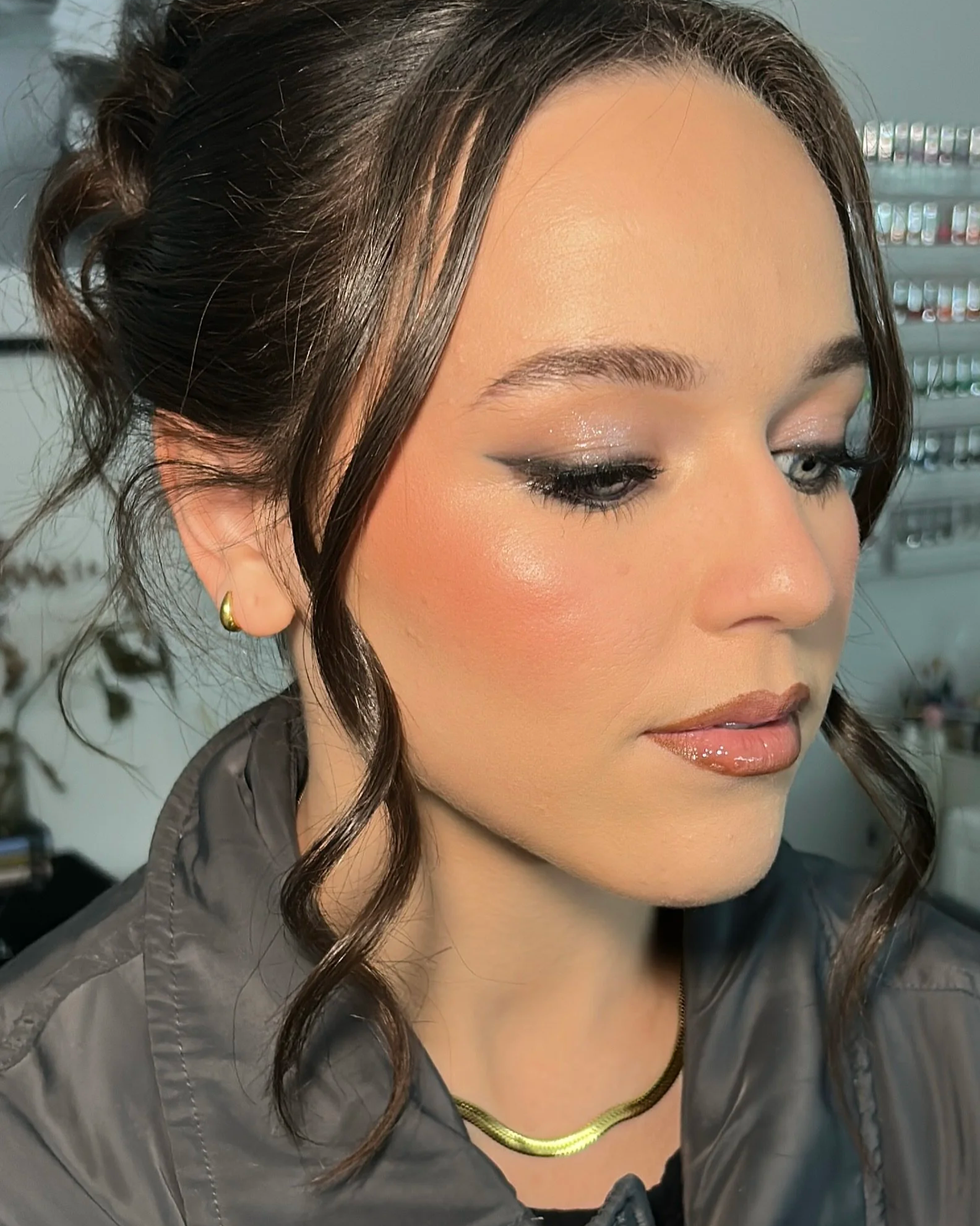 Close-up of a woman with styled dark brown hair, wearing makeup, a gold hoop earring, a gold necklace, and a gray jacket, indoors.
