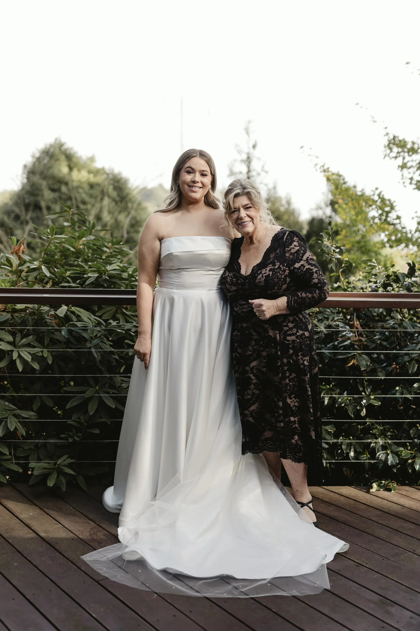 A young woman in a white wedding dress standing next to a woman in a black lace dress outdoors on a wooden deck with green trees in the background.