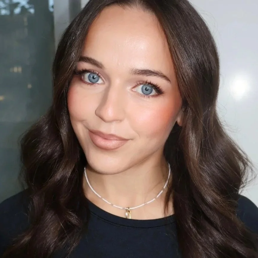 A close-up of a woman with long brown hair and blue eyes, looking directly at the camera with a neutral expression. She is wearing makeup, glossy lipstick, and a gold necklace.