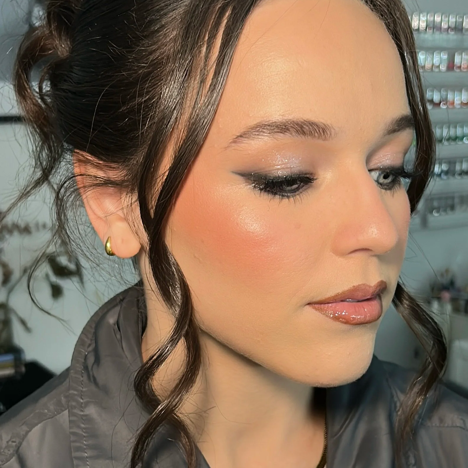 A close-up of a woman with long brown hair and blue eyes, looking directly at the camera with a neutral expression. She is wearing makeup, glossy lipstick, and a gold necklace.