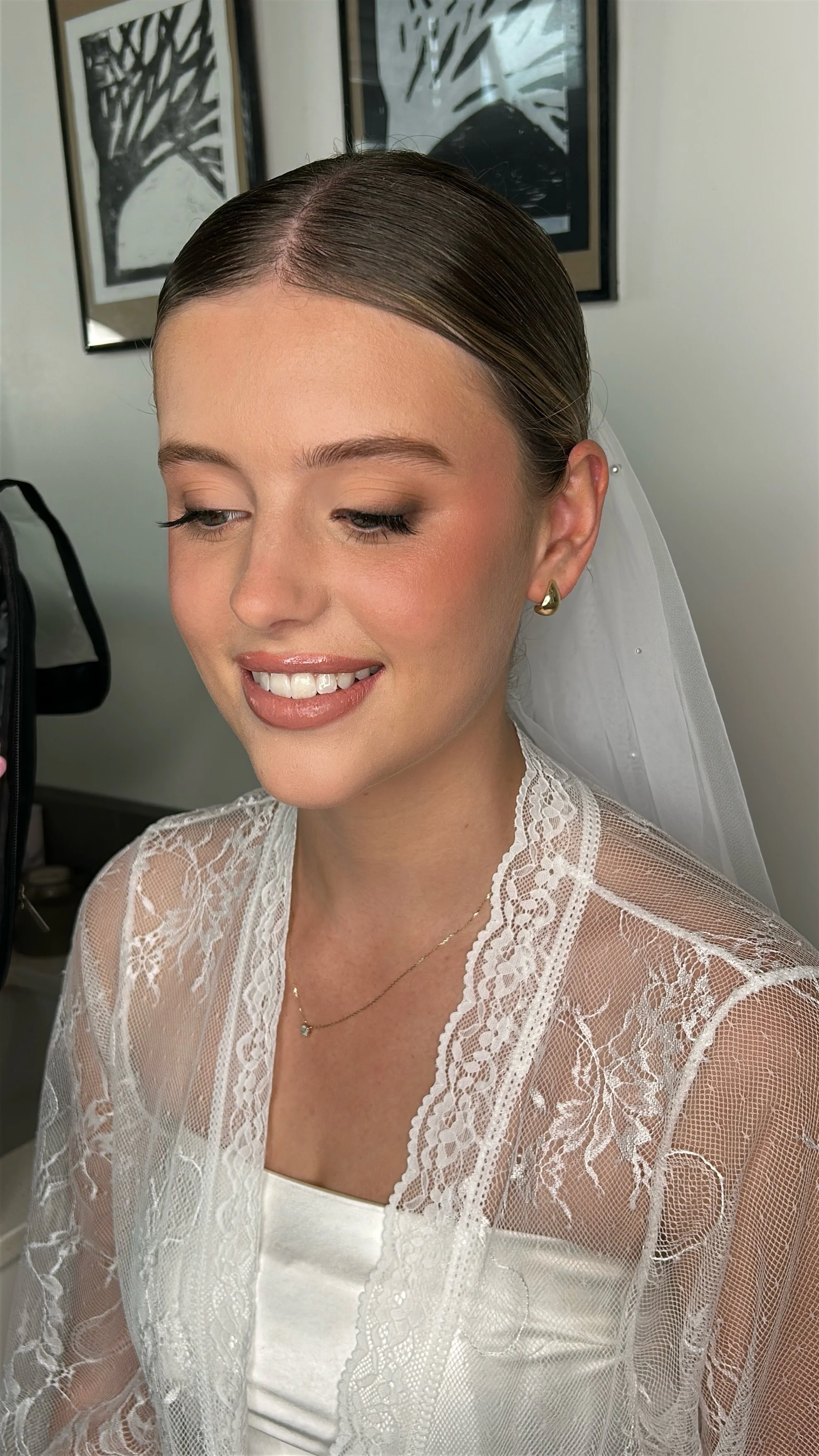 A bride with neat hair, makeup, and earrings in a satin dress with a lace overlay, smiling gently in a decorated room.