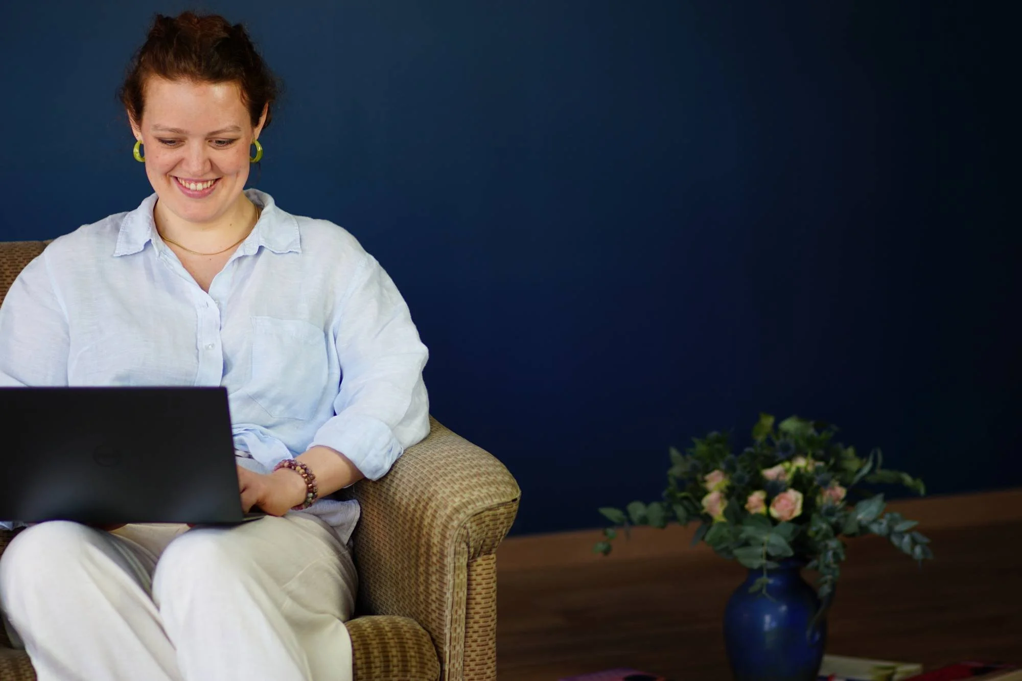 Eine lachende Frau sitzt auf einem gepolsterten Sessel und arbeitet an einem Laptop, im Hintergrund eine blaue Wand und eine Vase mit Blumen.