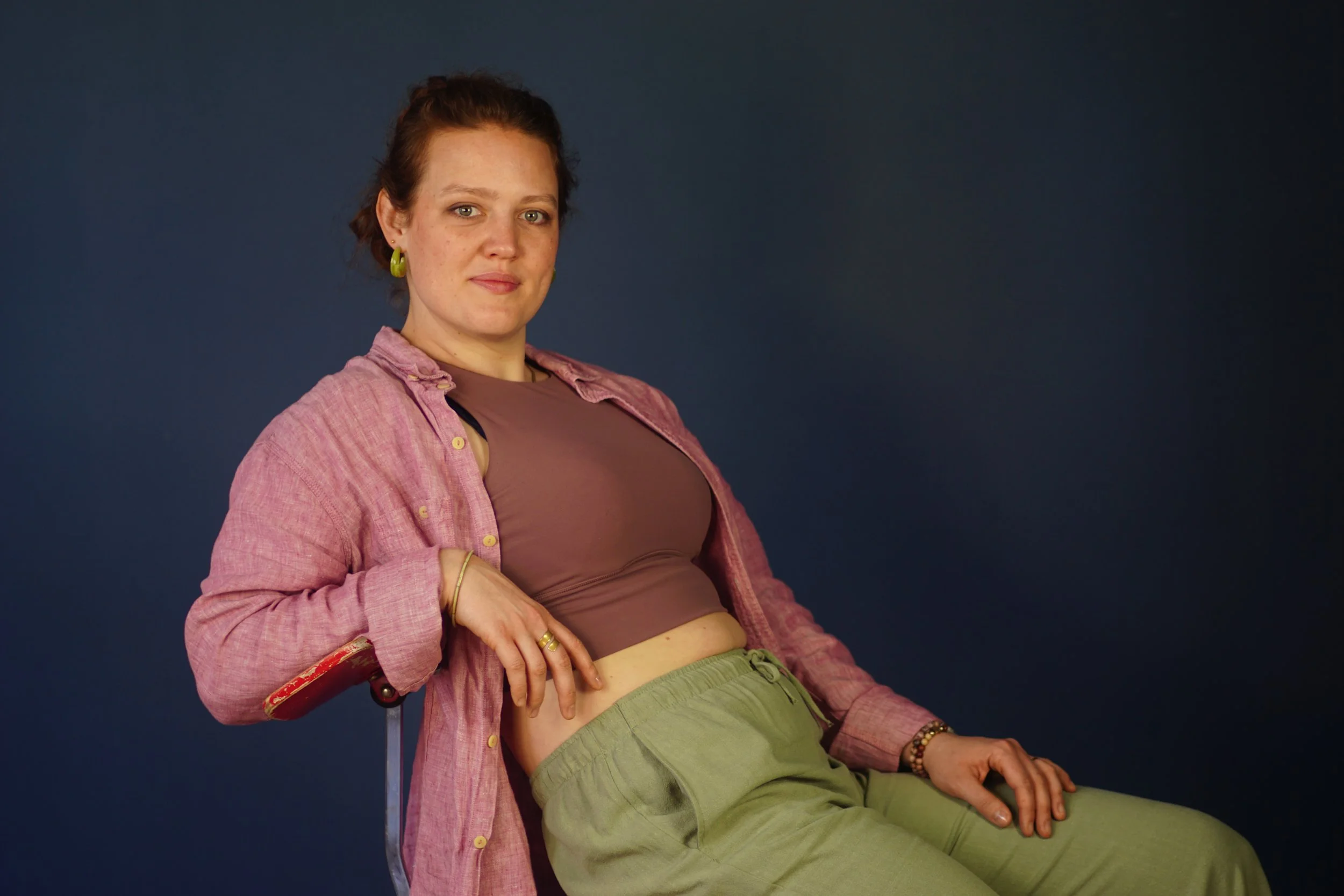 Junge Frau mit rotbraunen Haaren, lockig, sitzt auf einem Stuhl vor einem dunkelblauen Hintergrund, trägt ein braunes Top, eine offene rosafarbene Hemdjacke und grüne Jogginghose.
