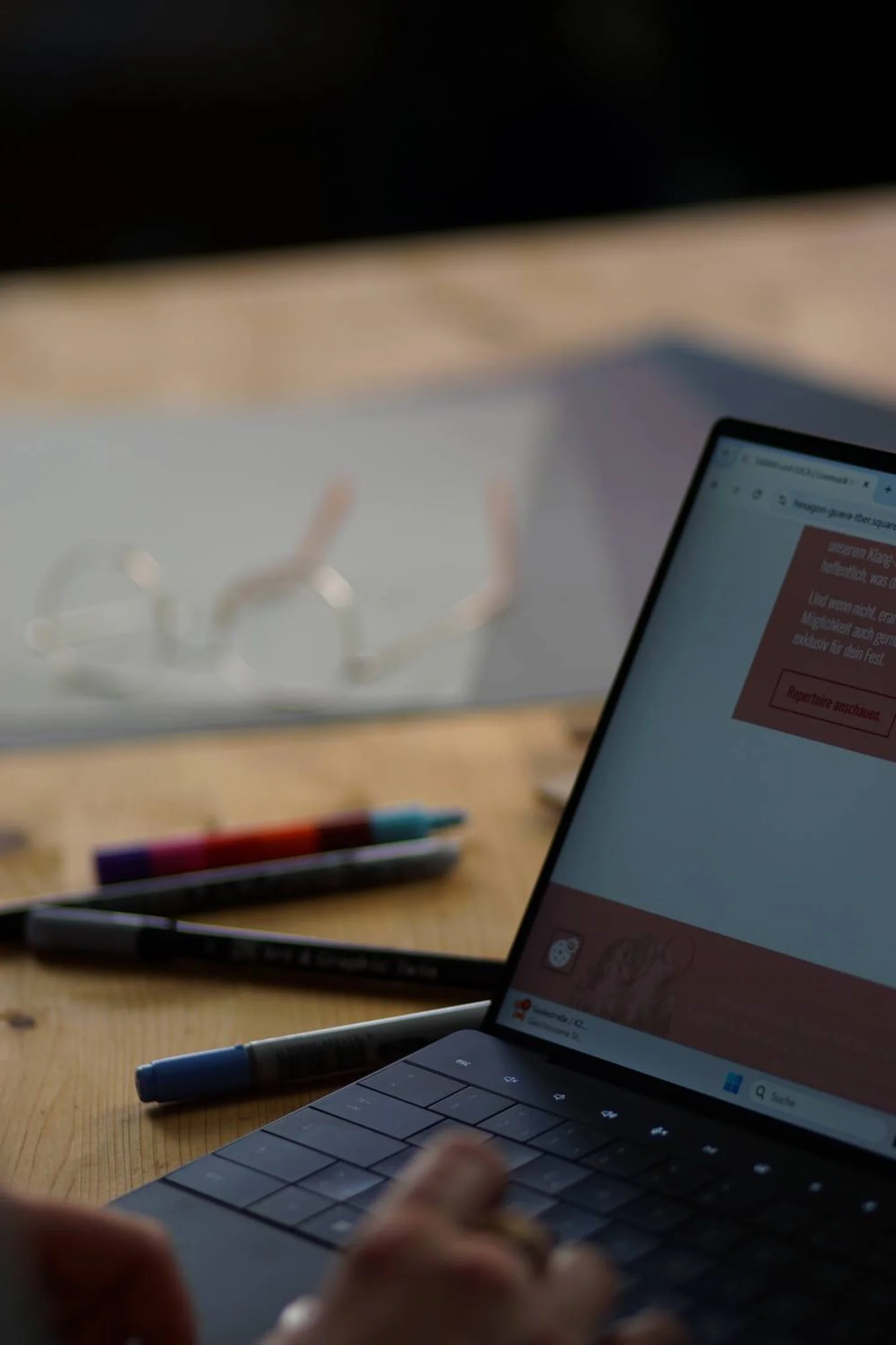 Nahaufnahme eines Laptops auf einem Holztisch mit einigen Filzstiften in der Nähe, im Hintergrund liegt eine Brille auf einem weißen Gegenstand. Der Bildschirm des Laptops zeigt eine Webseite in deutscher Sprache.
