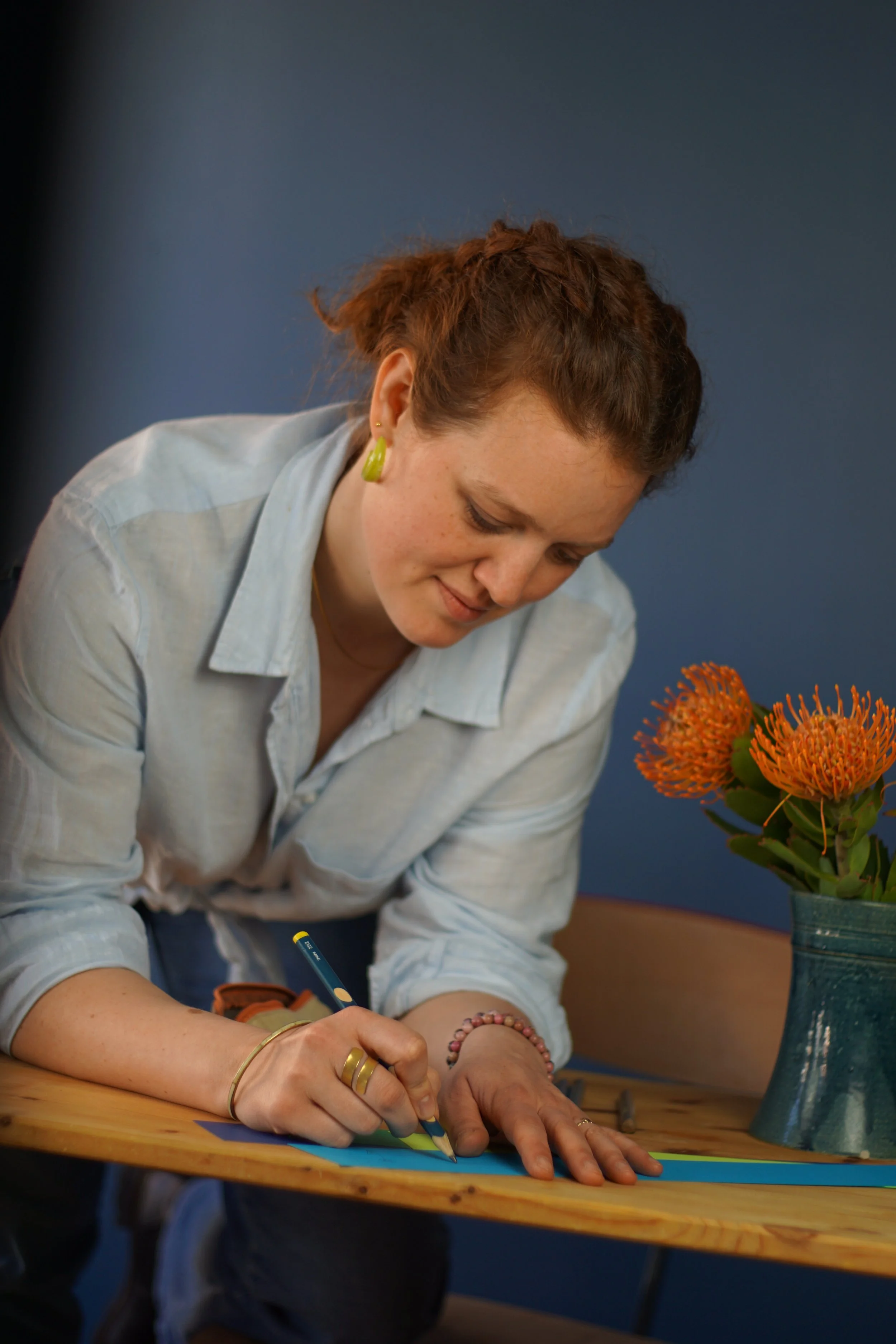 Eine Frau schreibt auf ein farbiges Papier auf einem Holztisch, neben einem Blumenstrauß mit orangefarbenen Blumen in einer blauen Vase.