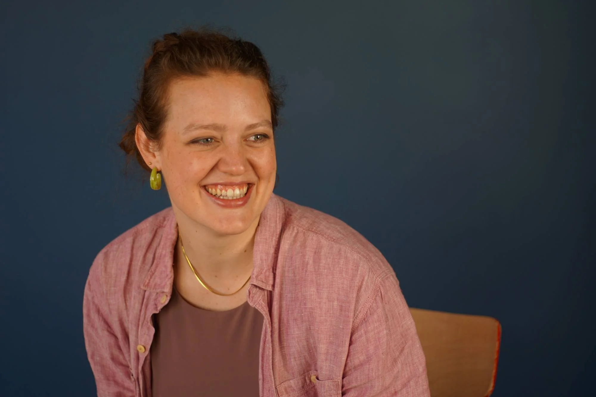 Lächelnde junge Frau, grünen Ohrringen, goldener Halskette, in rosa Hemd auf dunklem blauen Hintergrund.