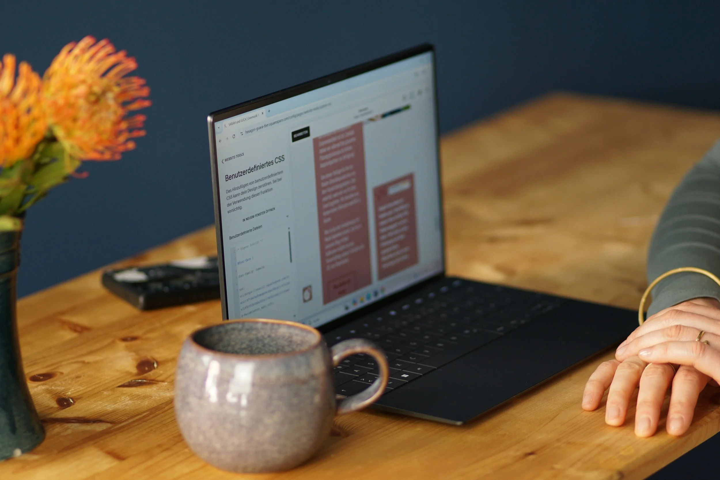 Ein Laptop auf einem Holztisch mit einer Person, die die rechte Hand auf dem Tisch hat. Es gibt eine Vase mit Blumen, eine Kaffeetasse, ein Handy und einen blauen Wandhintergrund.
