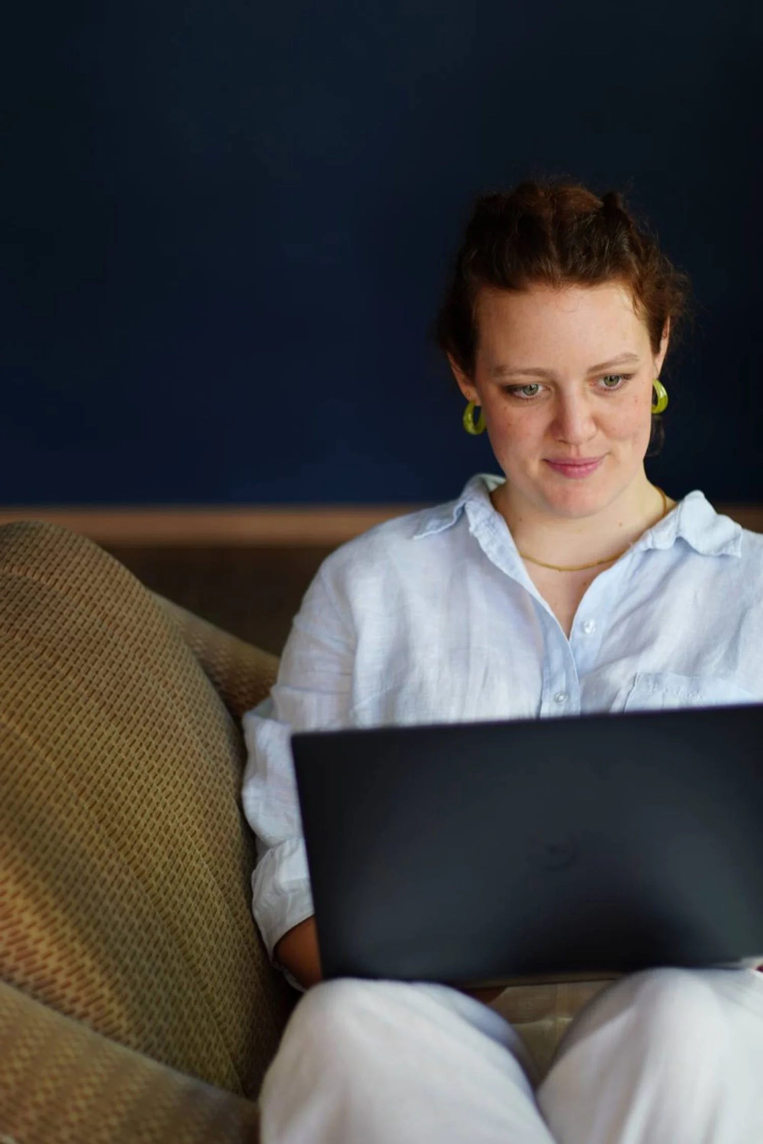 Junge Frau mit roten Haaren und grünen Ohrringen sitzt auf einem Sitz und arbeitet an einem Laptop.