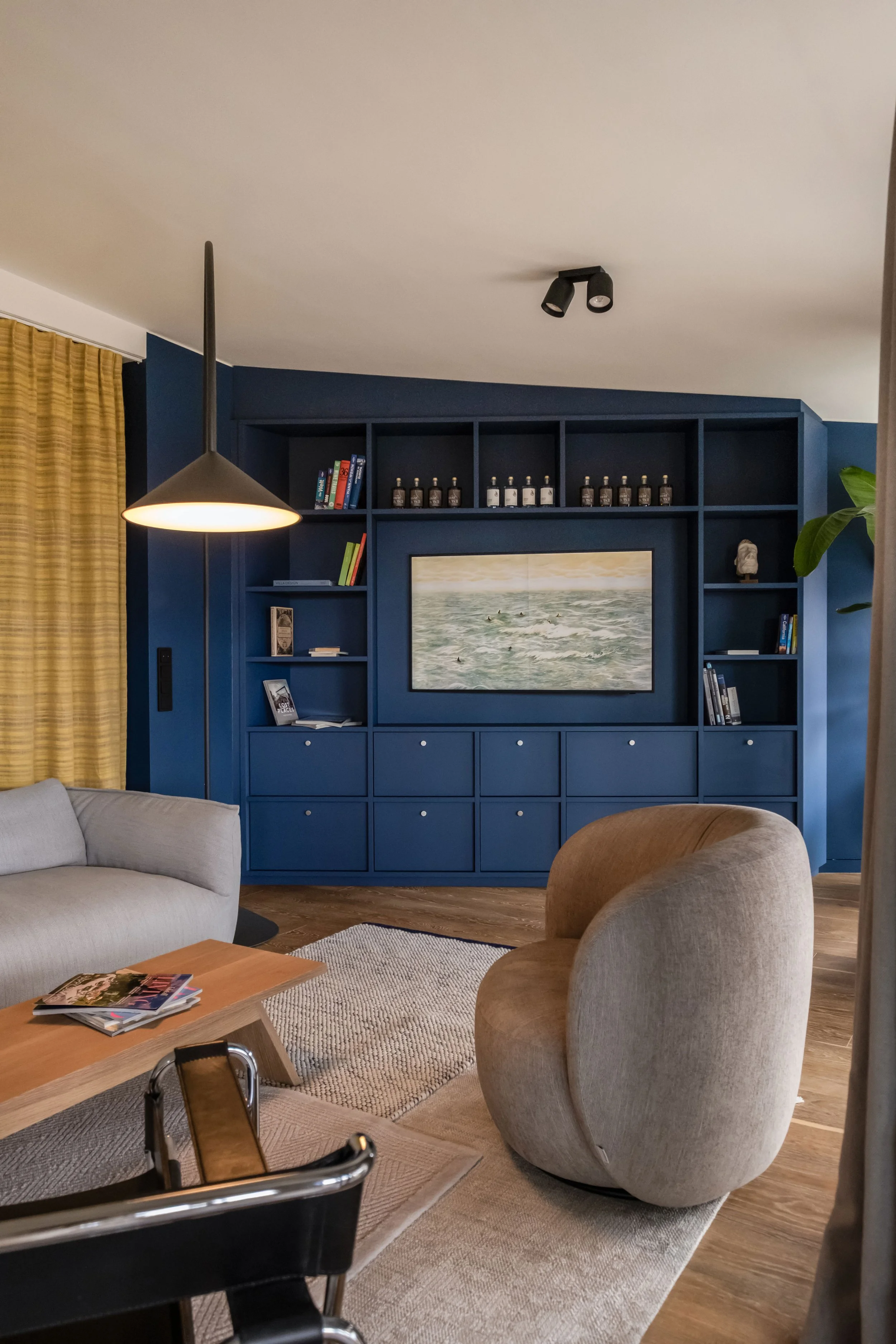 Ein Wohnzimmer mit blauer Wand, einem Sofa, einem Sessel, einem Couchtisch mit Magazinen, einer Stehlampe, einer Bücherwand und einem TV mit einem Gemälde mit Wellen im Meer.