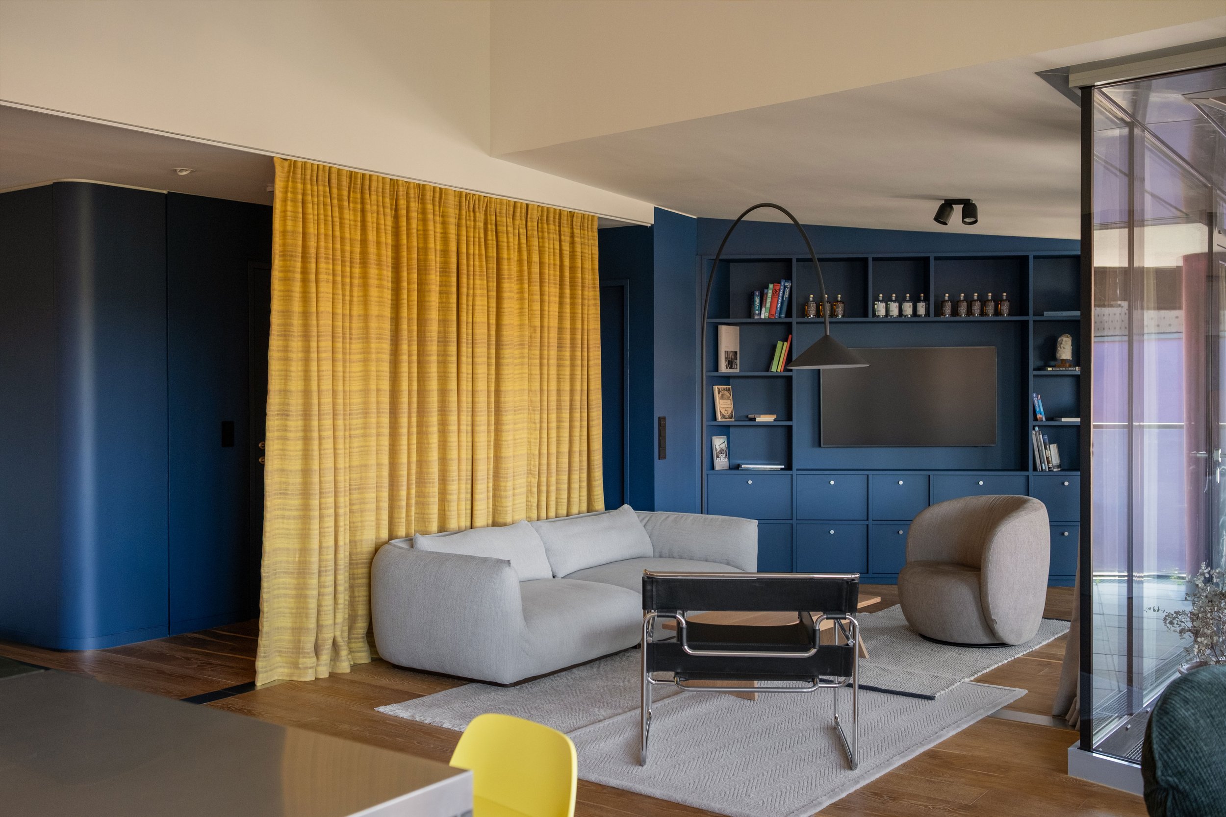 Ein Wohnzimmer mit dunkelblauen Wänden, gelben Vorhängen, einem hellblauen Sofa, einem beigen Sessel, einem Wassily Chair und hellen Teppichen. Es gibt ein Bücherregal mit Büchern und Dekorationsgegenständen und einen Lampenschirm über dem Couchtisch