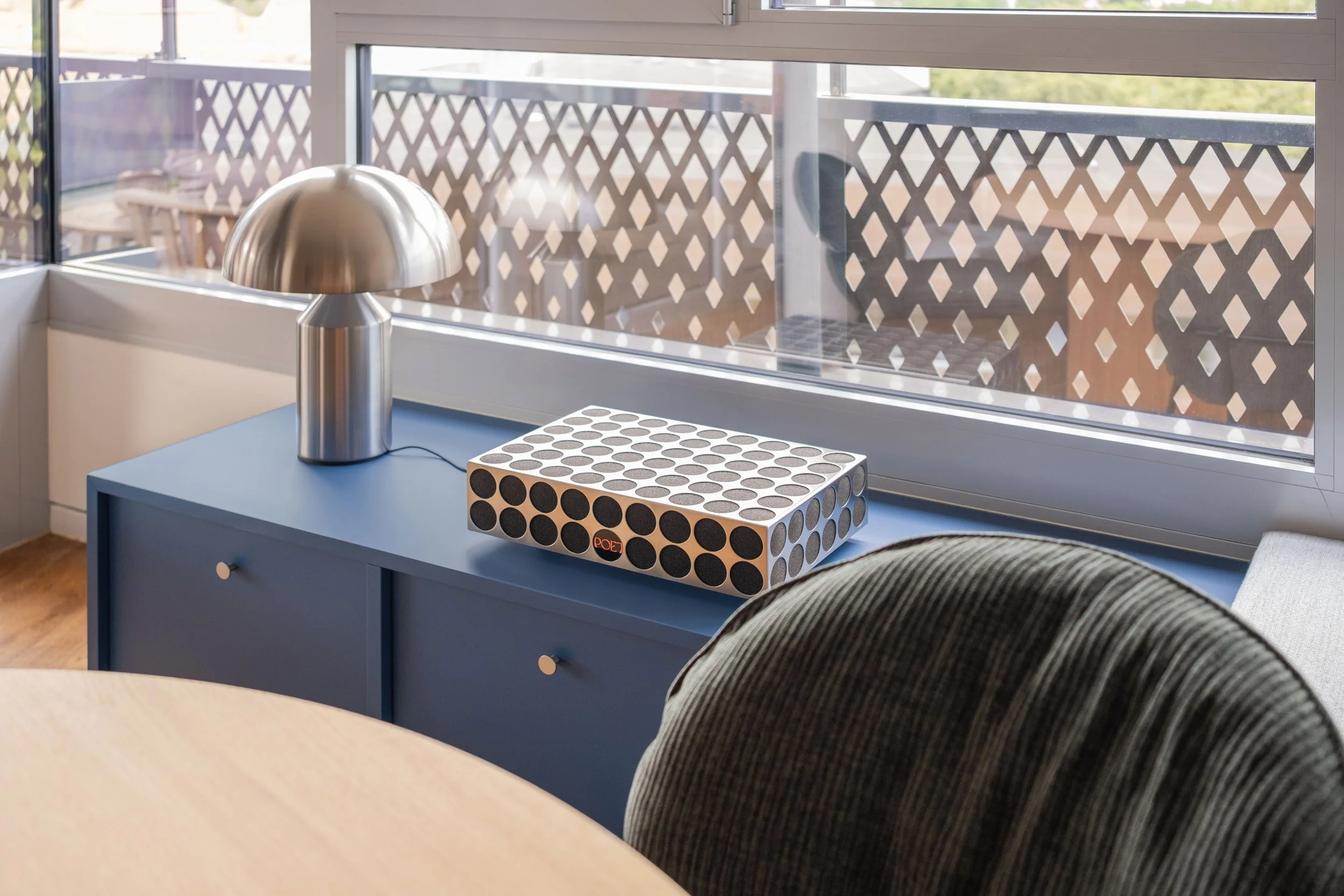 Ein blaues Sideboard mit einer modernen Tischlampe aus Edelstahl, einer Soundanlage vor einem Fenster.