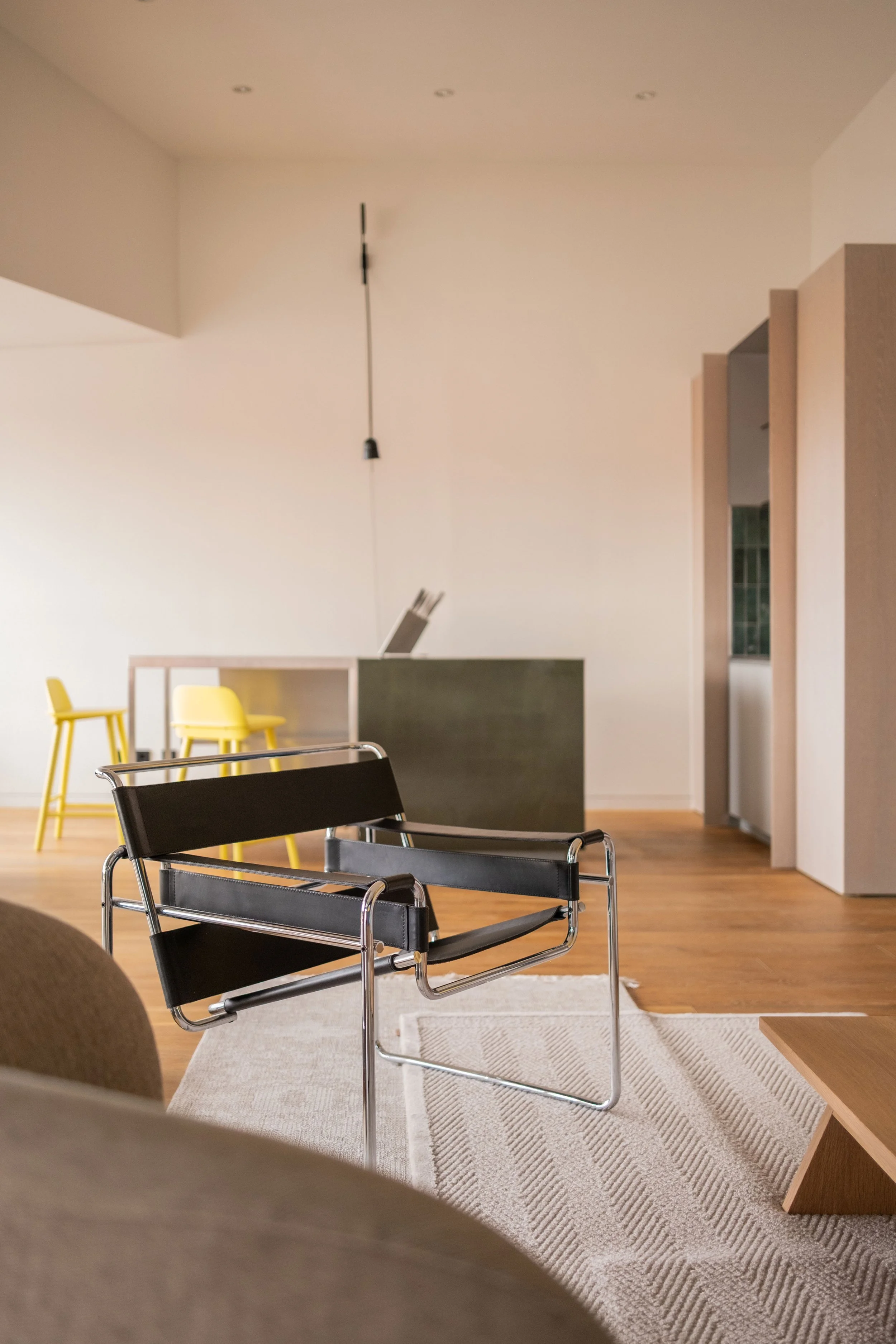 Modernes Wohnzimmer mit schwarzen Loungesessel, hellem Teppich, im Hintergrund eine Kücheninsel mit gelben Hockern, offener Grundriss, helle Wandfarben, Holzboden.