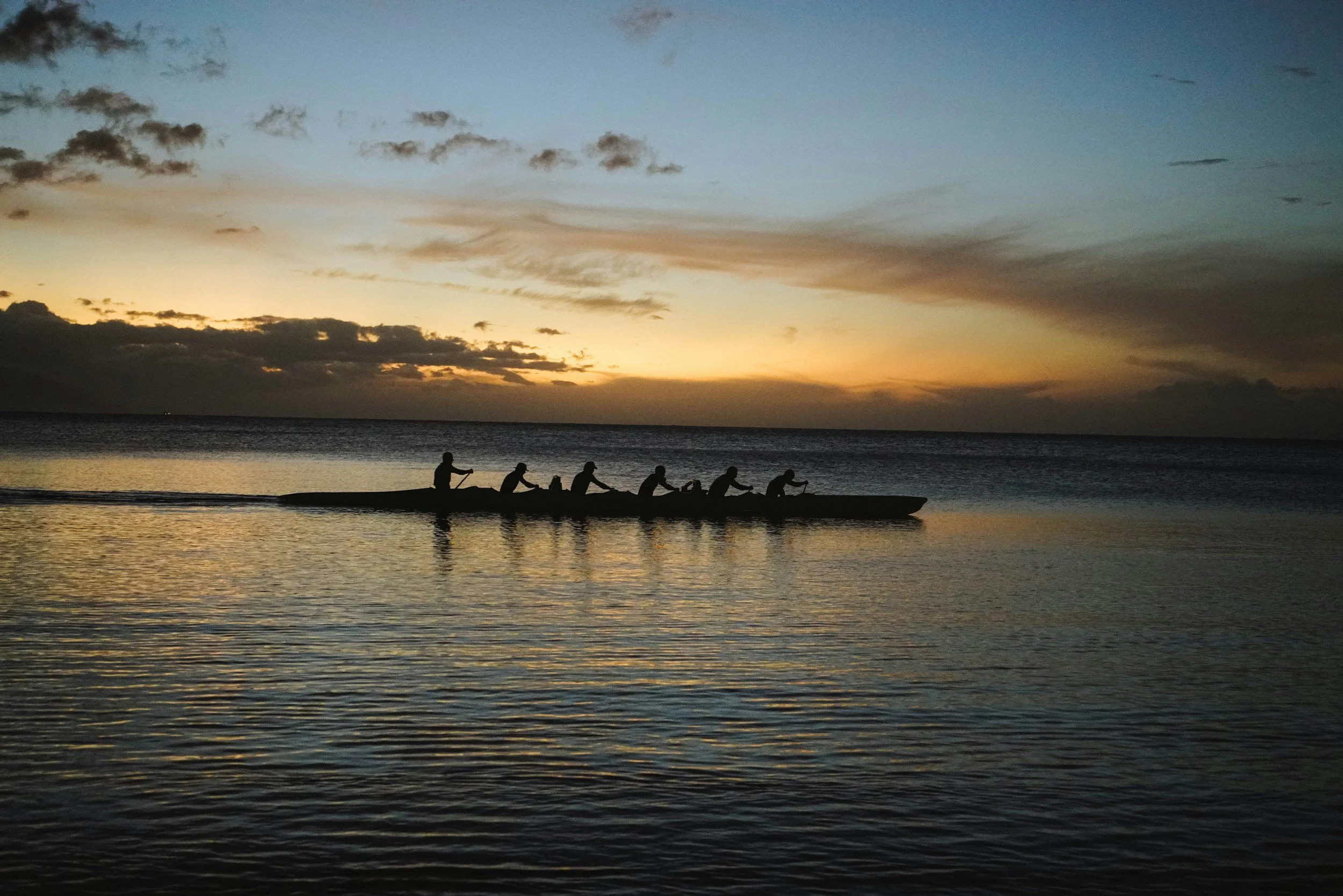 Silhouet van zes mensen in een boot, roeien op een kalm meer tijdens zonsondergang, met een kleurrijke hemel en bewolking.