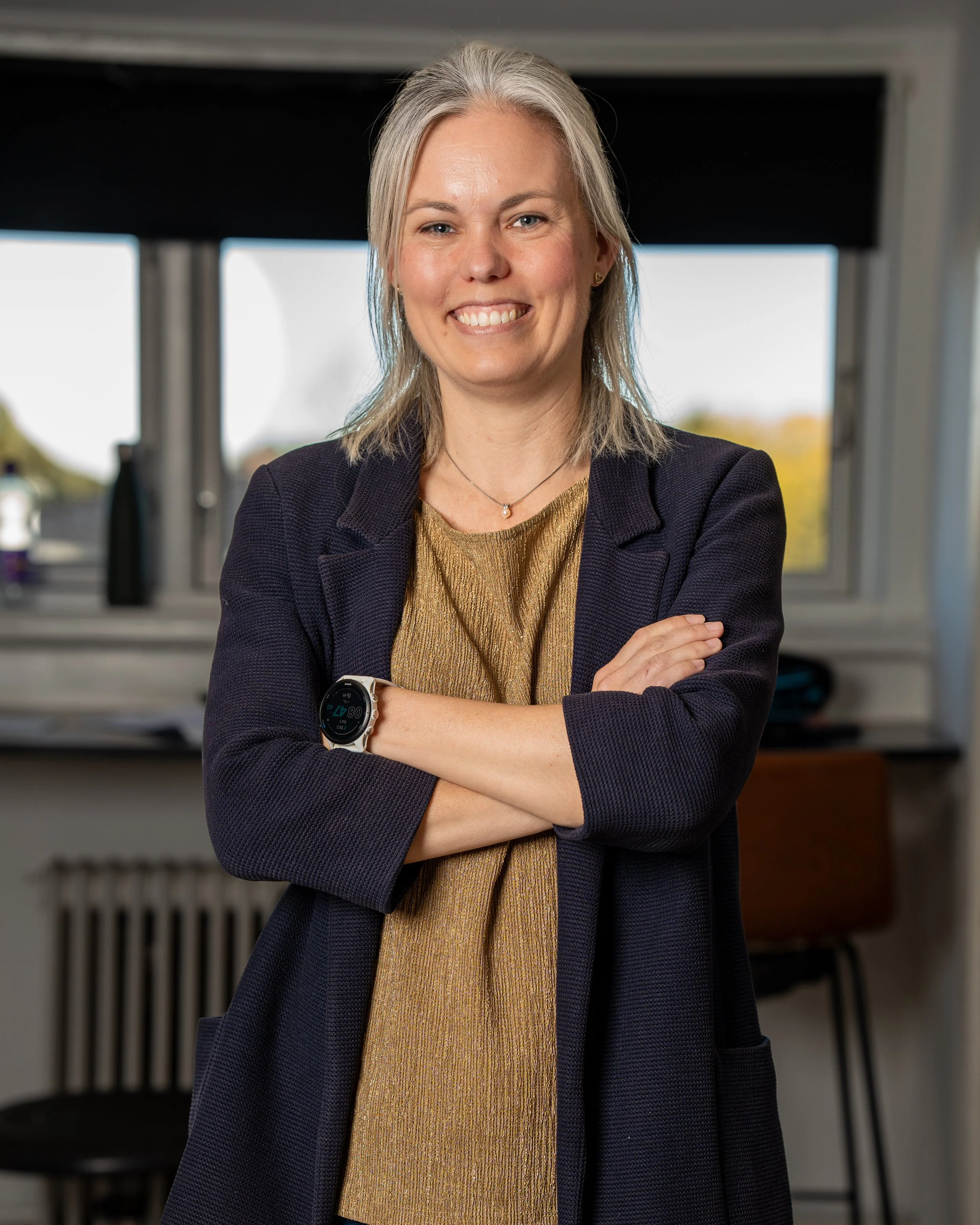 En smilende kvinde med gråt hår, iført en guld top og en mørk blazer, står med armene krydset i en moderne indendørs setting.
