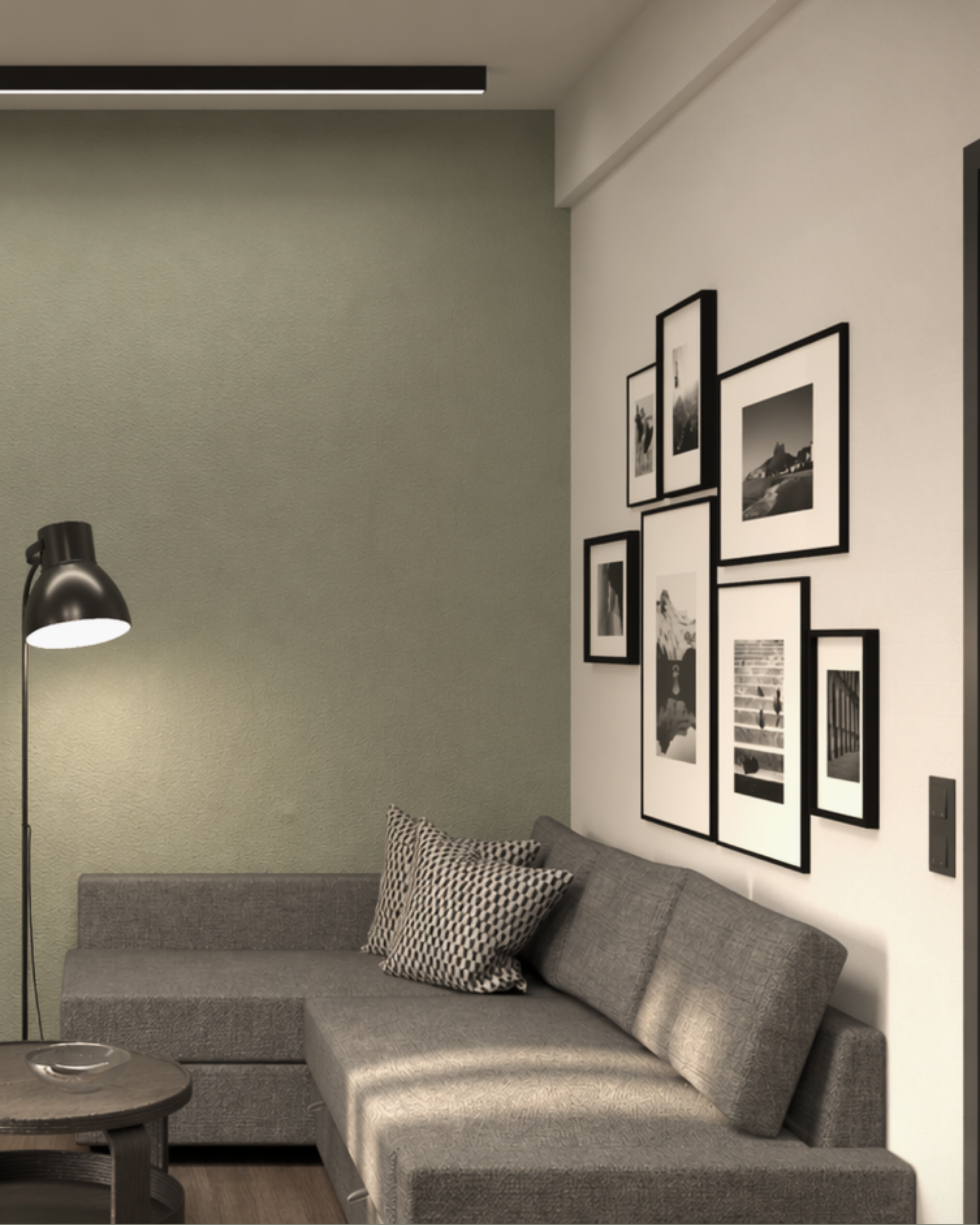 A living room corner with a gray sectional sofa adorned with patterned pillows, a small round side table, a black adjustable lamp, a gallery wall with black and white framed photographs, a green accent wall, and a ceiling light.
