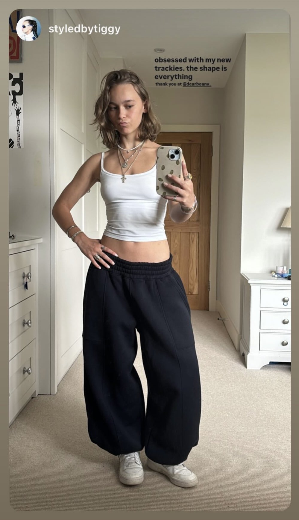 A young woman taking a mirror selfie in a bedroom, wearing a white cropped tank top, black wide-leg pants, and white sneakers. She has shoulder-length wavy hair, multiple necklaces, and rings.