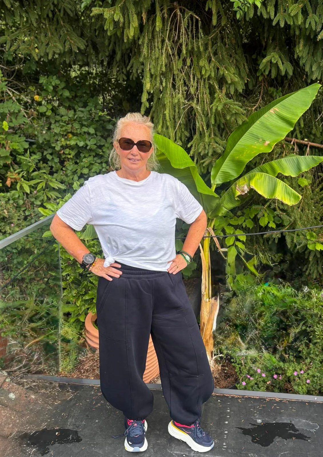 A woman in sunglasses standing with her hands on her hips in front of lush green tropical plants, including large banana leaves and other foliage.