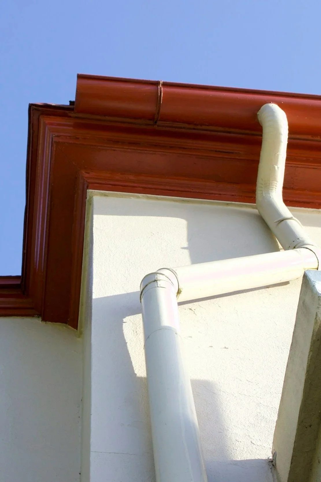 Close-up view of a corner of a building with a red roof, white wall, and white drainage pipes against a clear blue sky.
