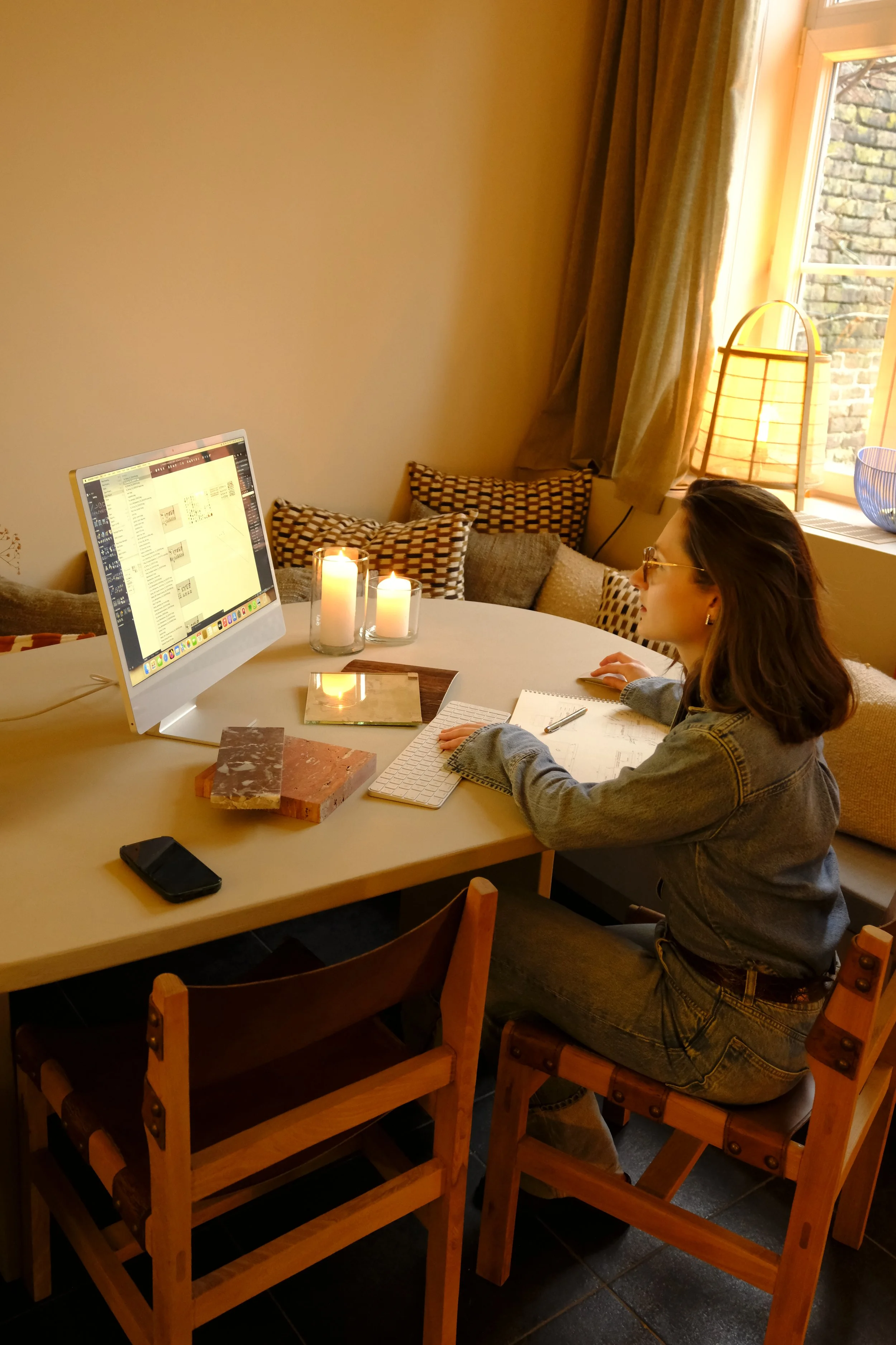 A woman working at a desk with a computer, candles, notebooks, and a pen in a cozy, well-lit room.
