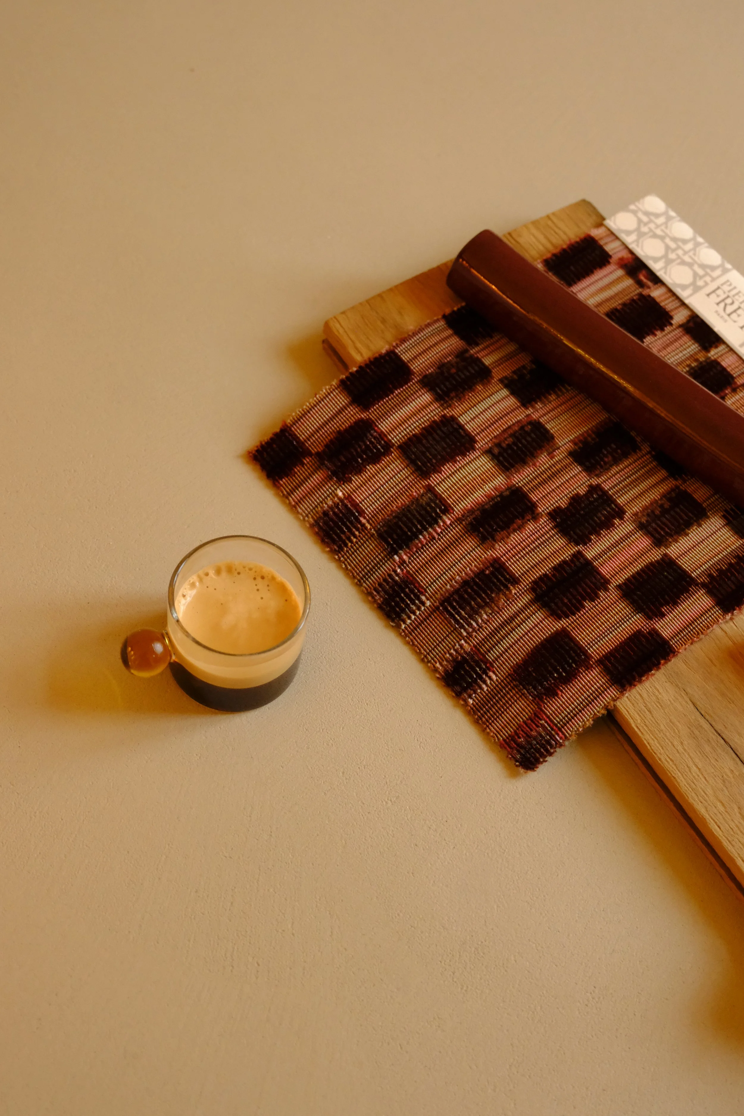 A glass of dark beer with a tan head on a light surface, next to a woven placemat and wooden tray with a paper napkin on top.