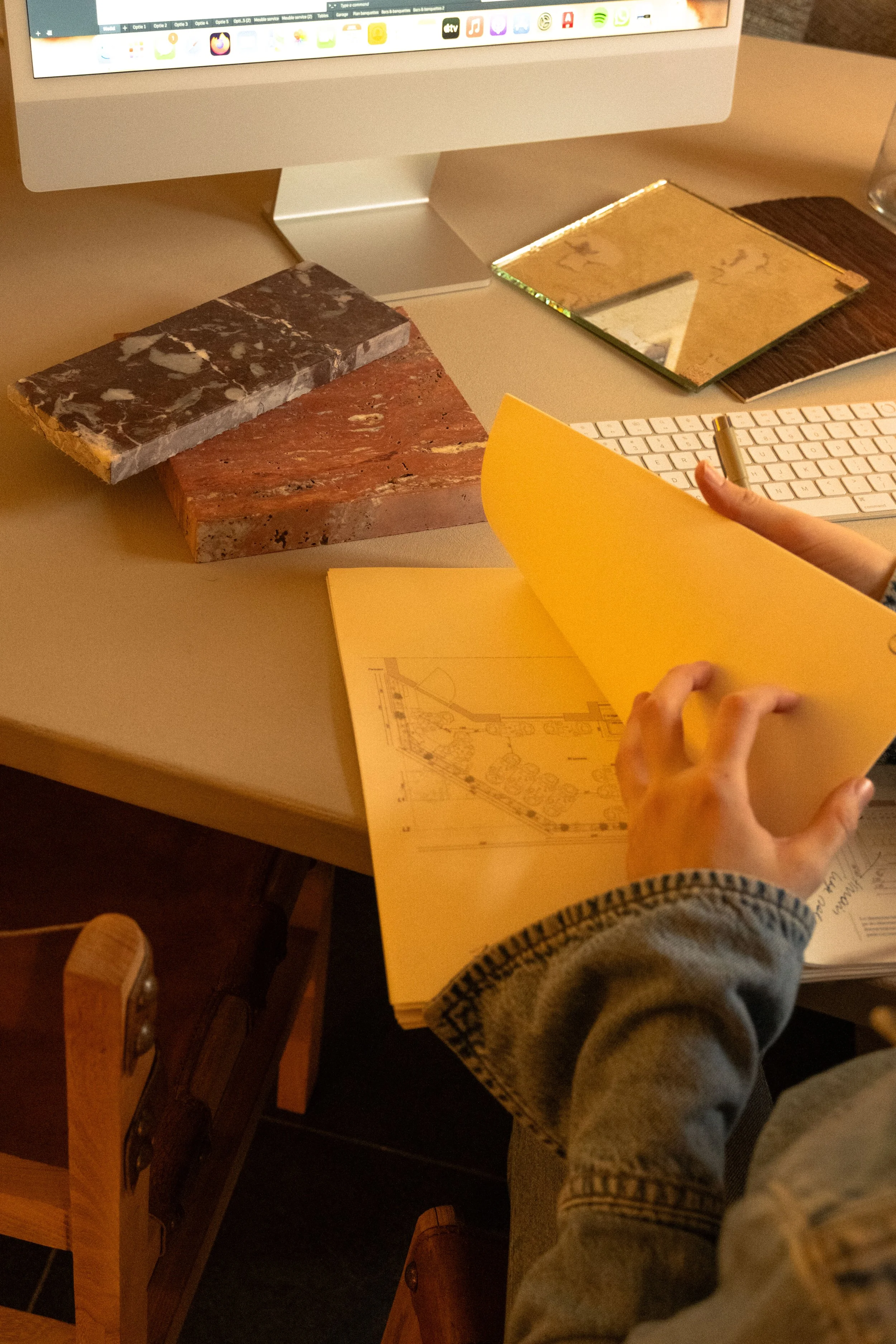 A person's hands flipping through a yellow sketchbook on a desk, surrounded by marble samples, a computer monitor, a white keyboard, and a mirror.