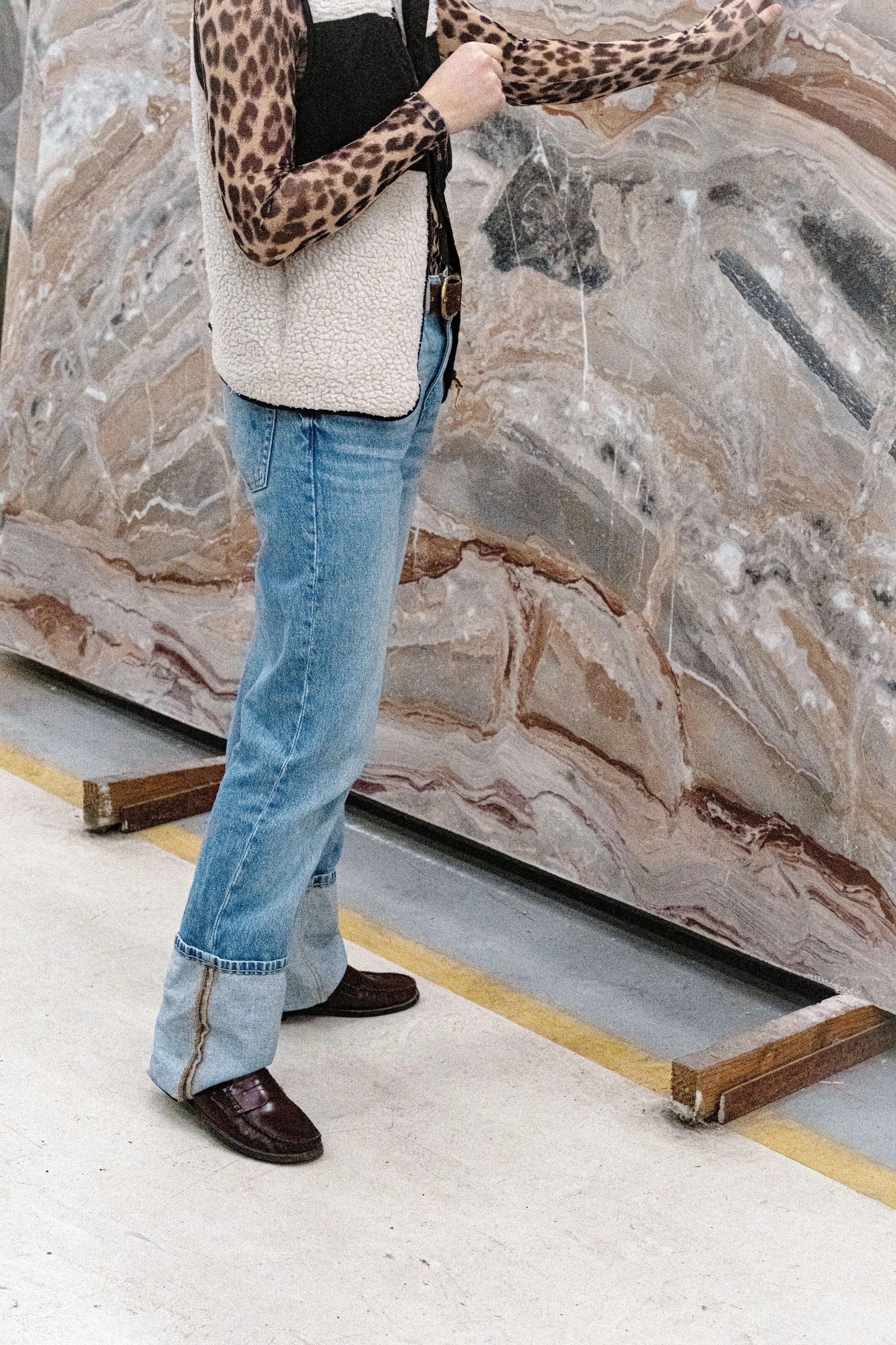 Person standing next to a large marble wall, wearing a leopard print long sleeve shirt, blue jeans, and brown shoes, carrying a cream-colored purse with a black strap.