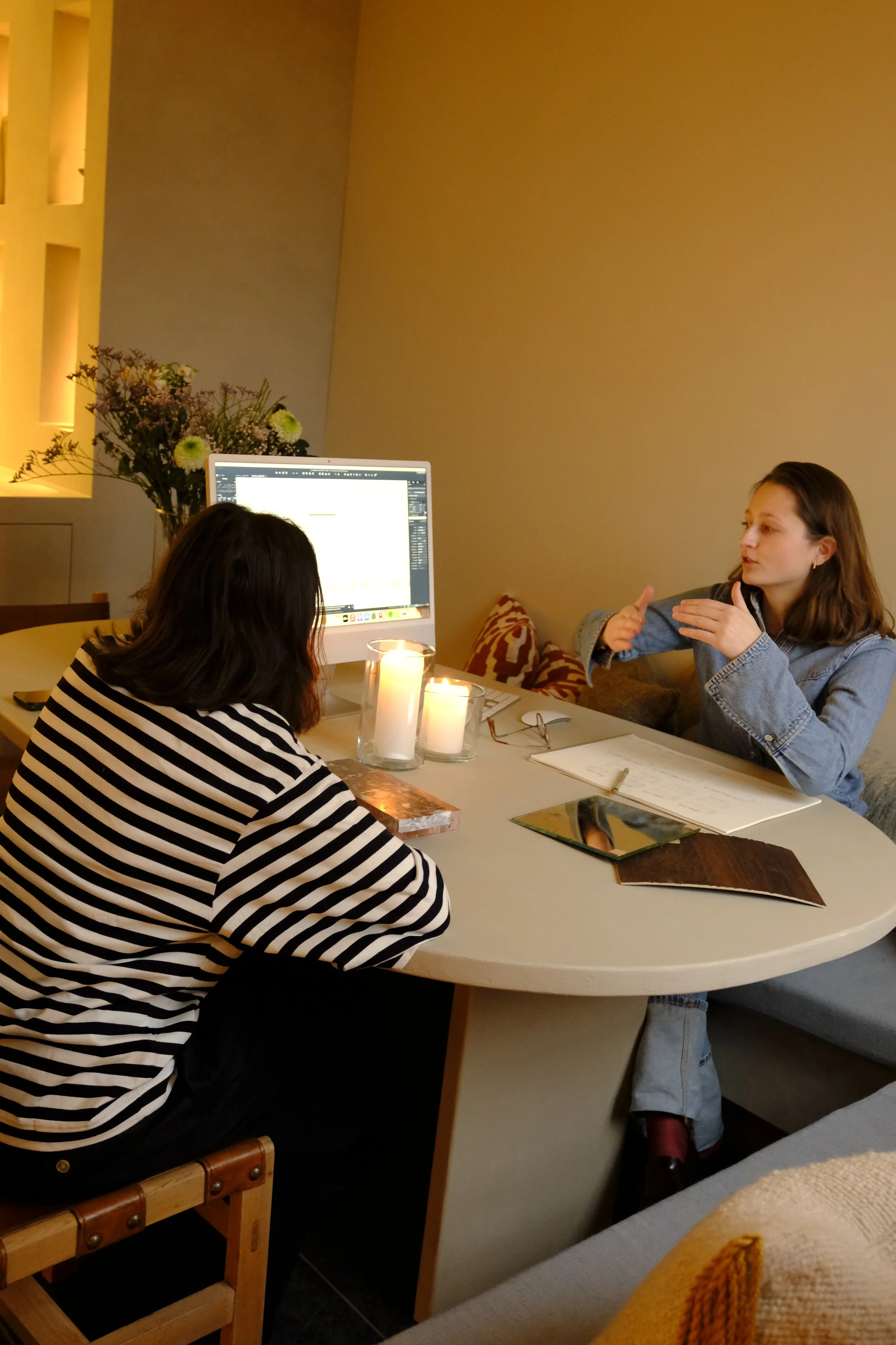 Two women having a discussion at a round table with candles, a computer monitor, notebooks, and folders in a cozy room