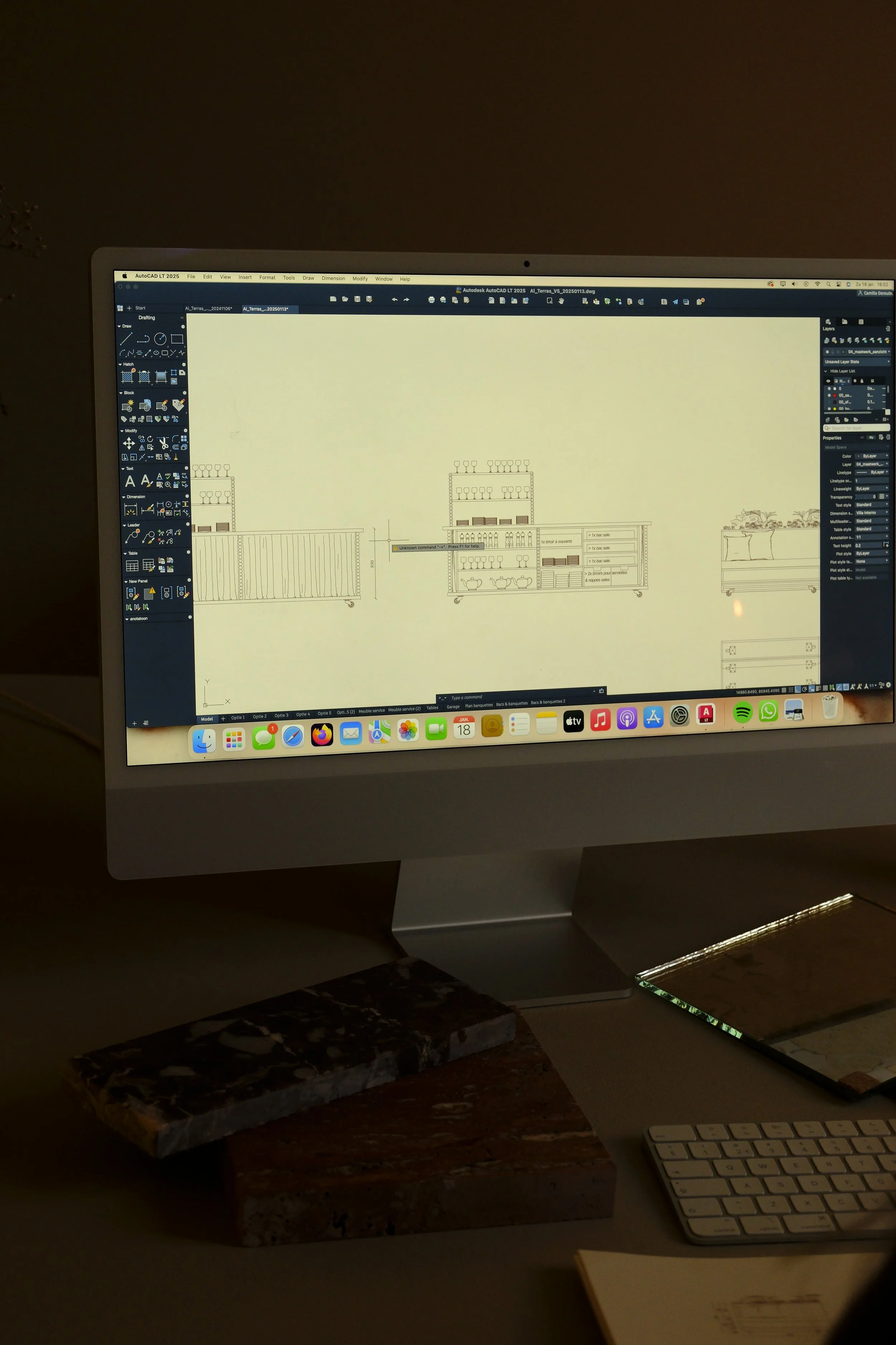 A computer screen displaying architectural design software with a technical drawing of shelves. The desk has two marble pieces, a wireless keyboard, a notebook, and a MacBook.