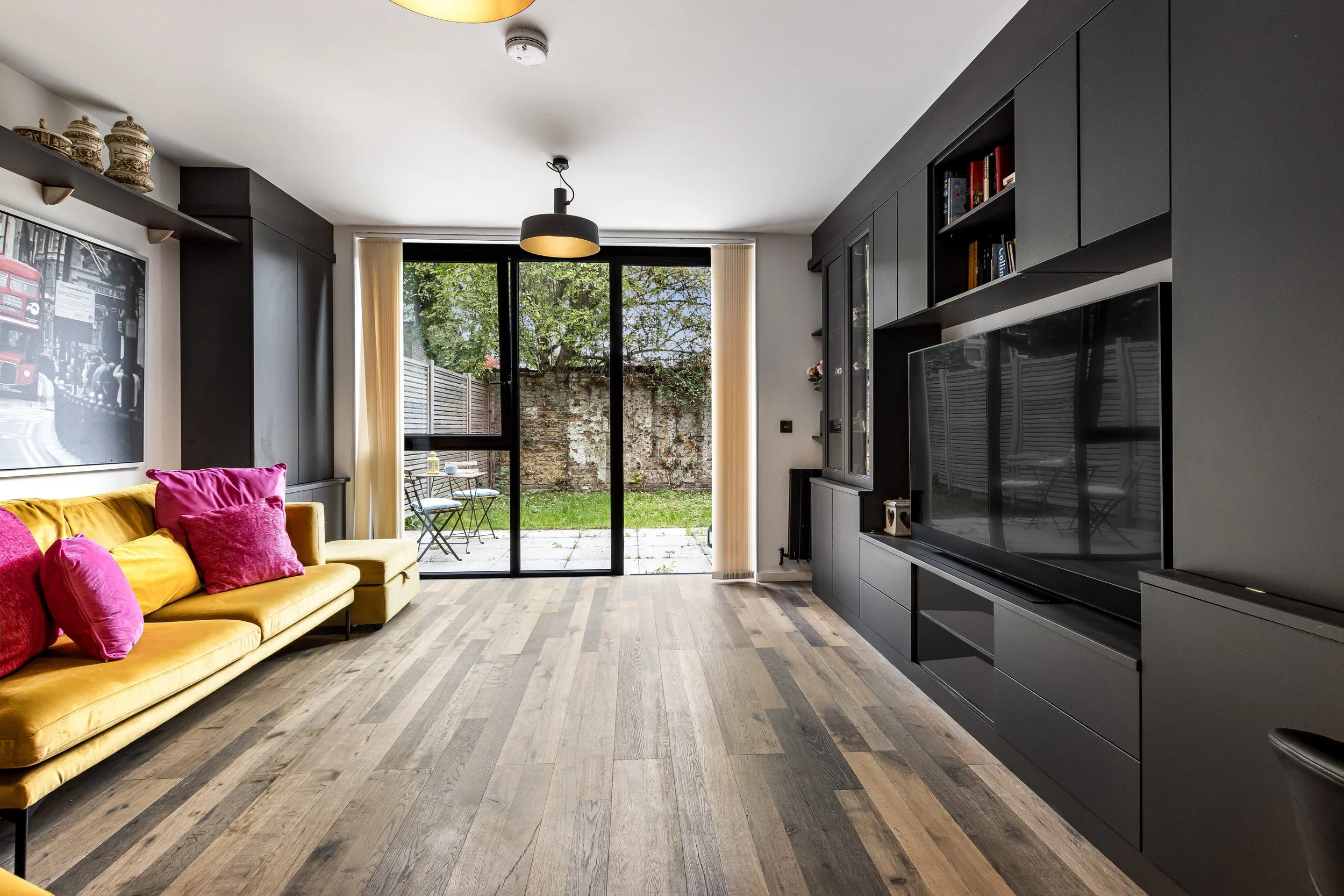 Modern living room with black built-in cabinets, a large flatscreen TV, a mustard-colored couch with pink and yellow cushions, and a view of a garden outside through glass sliding doors.