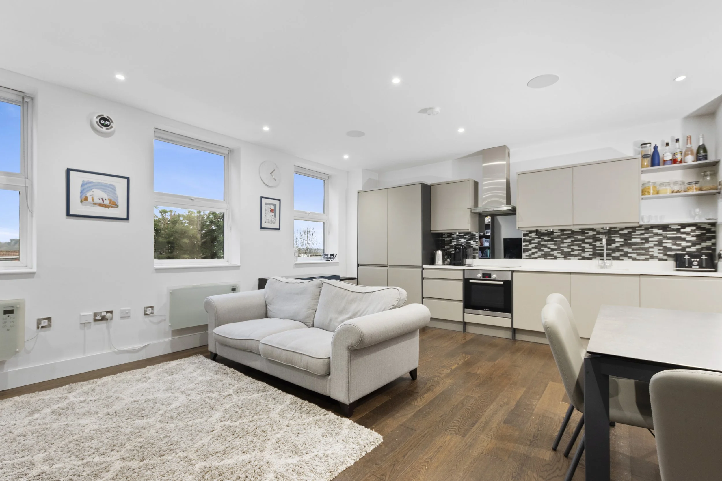 Modern living room with white walls, three large windows, a beige couch, a white shaggy rug, and a kitchen area with beige cabinets and a black mosaic tile backsplash.