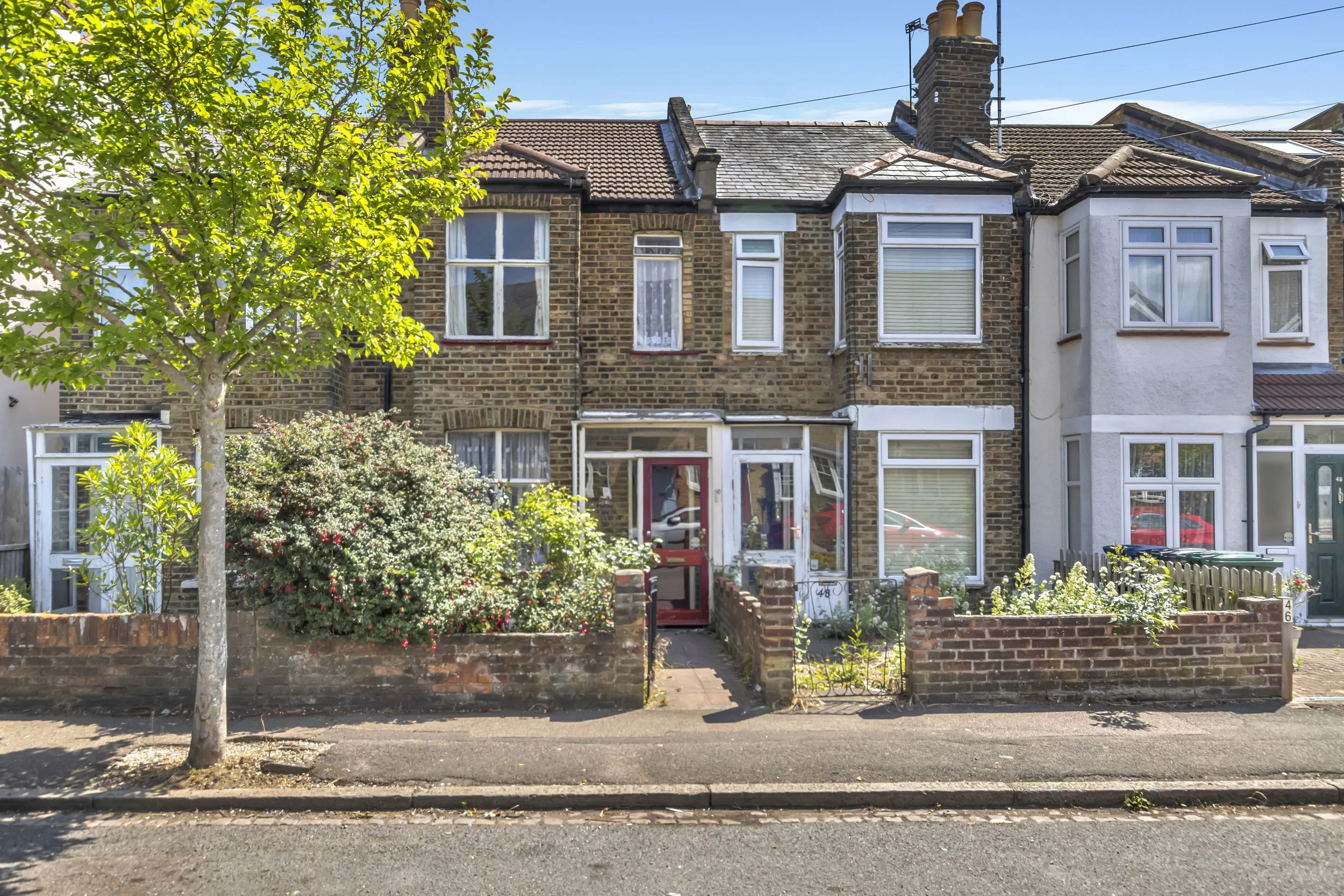 A row of residential houses with brick and white exteriors, front gardens, and trees along the pavement on a sunny day.