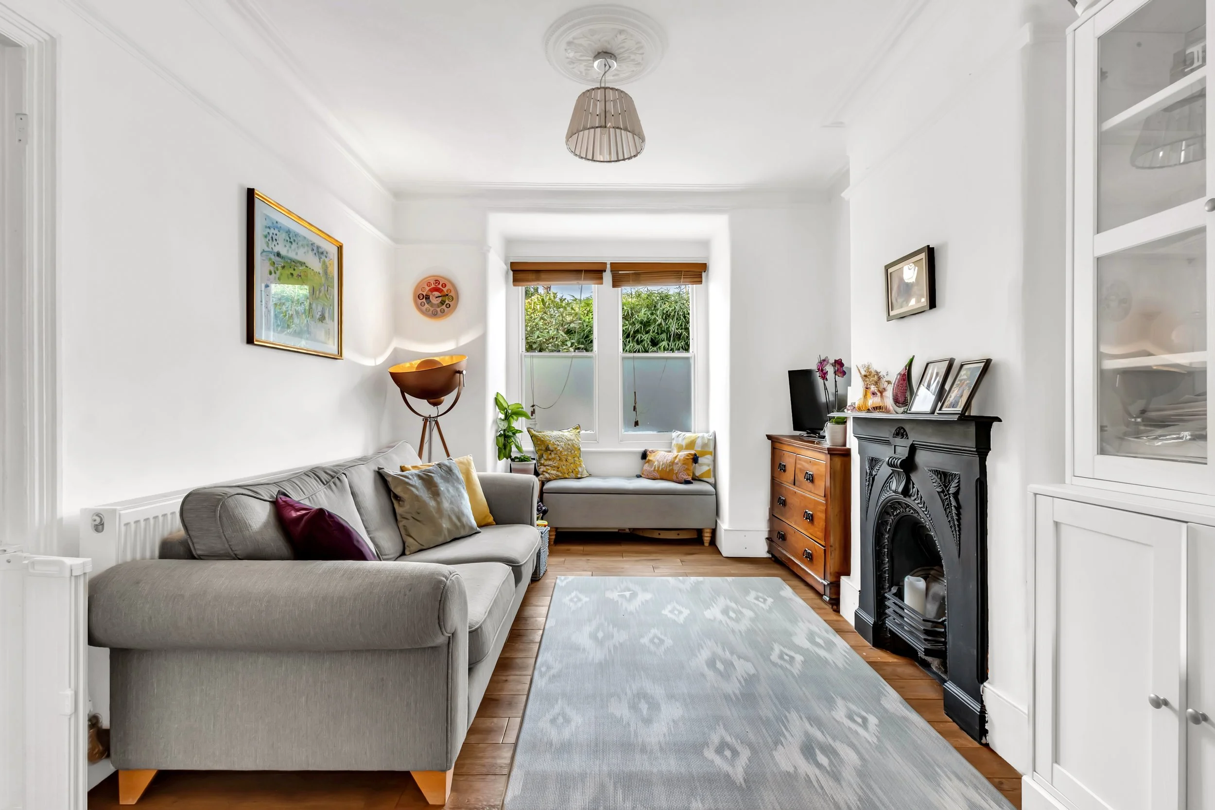 Living room with a gray sofa, a window seat with pillows, a black fireplace, a wooden chest of drawers, a small TV, and decorative items, with white walls and hardwood floors.