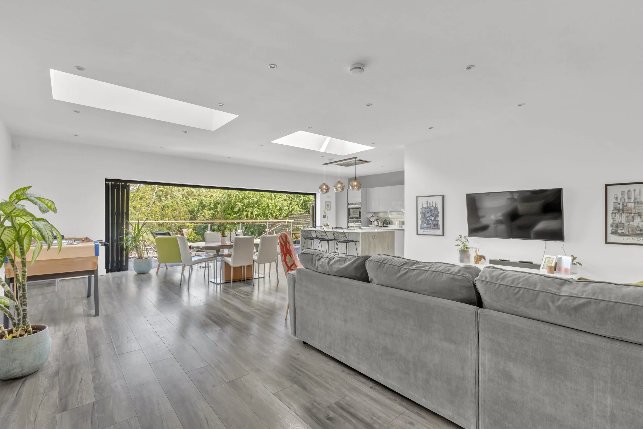 Modern open-concept living room with gray sofa, plants, dining area near large sliding doors, and kitchen with pendant lights.