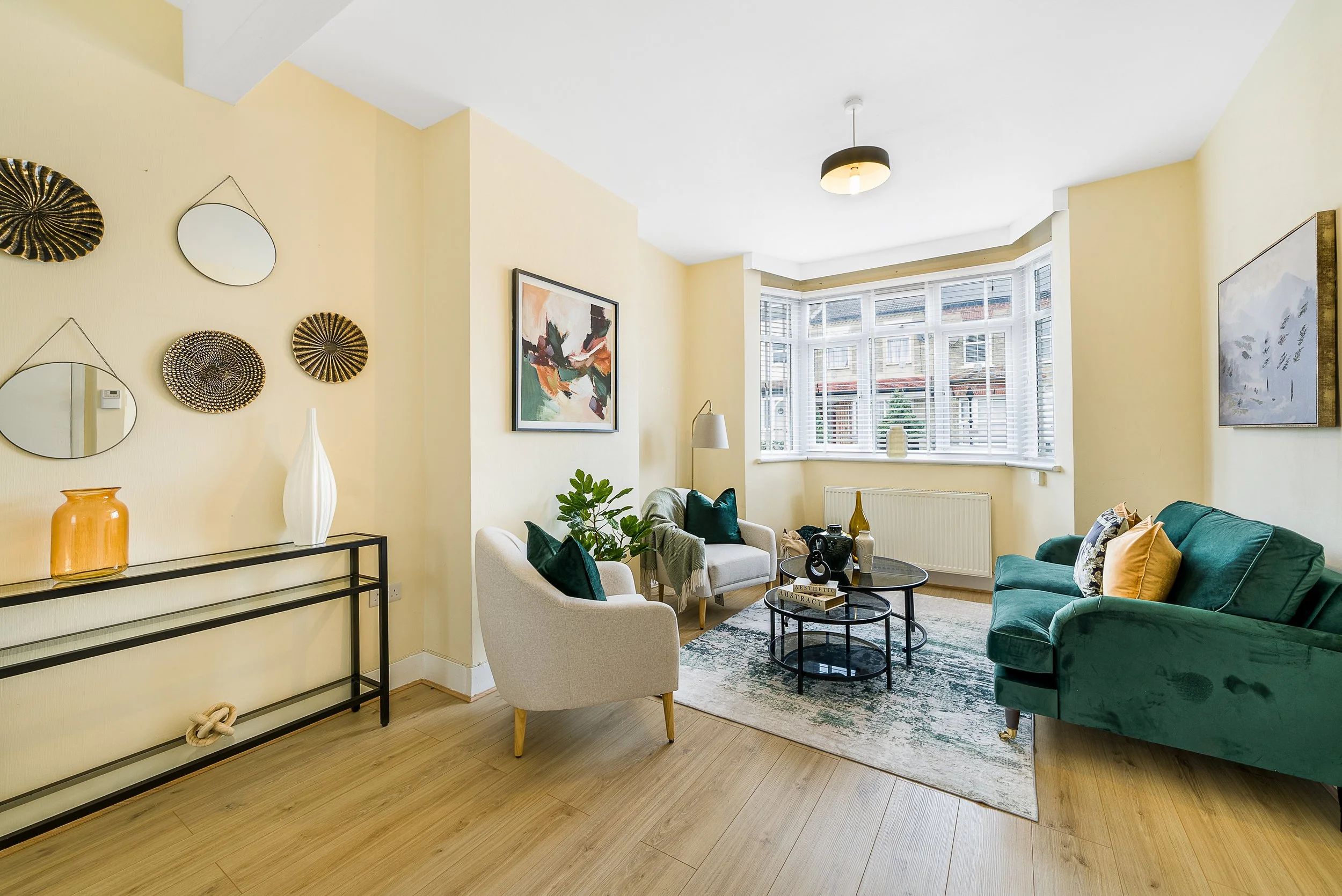 Living room with a large bay window, green velvet sofa with pillows, white armchair with pillows, and a black coffee table. Decor includes artwork, mirrors, and vases. Light-colored walls and wooden flooring.